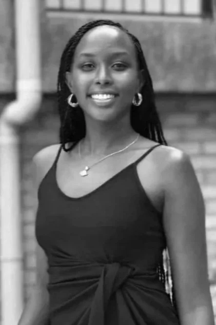 Smiling woman with long hair wearing a sleeveless dress, earrings, and necklace, standing outdoors in front of a brick building.
