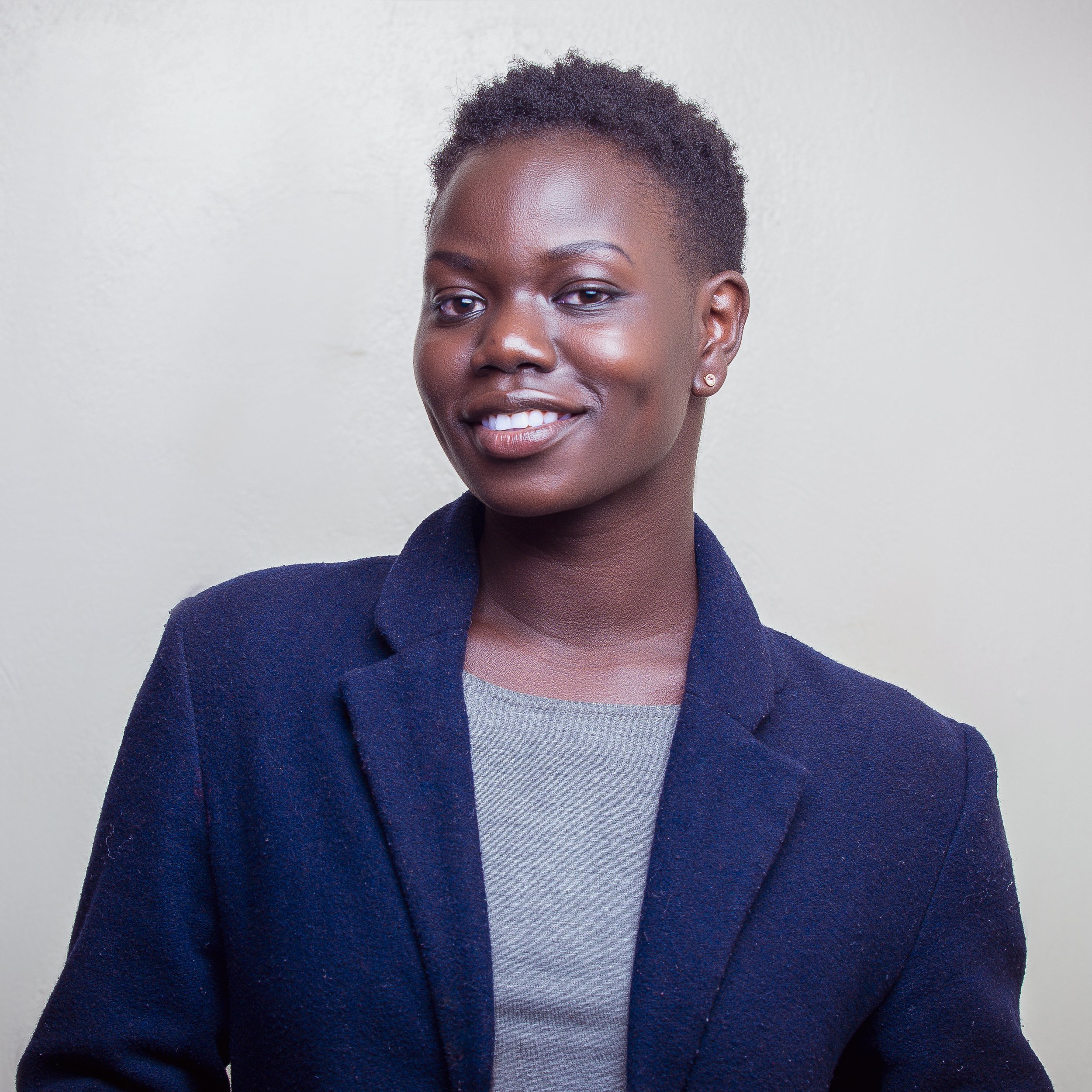 Portrait of a woman with short natural hair, wearing a dark blazer and a light gray top, smiling against a plain background.