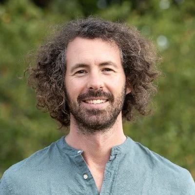 A man with curly hair and a beard smiling outdoors, wearing a blue shirt.