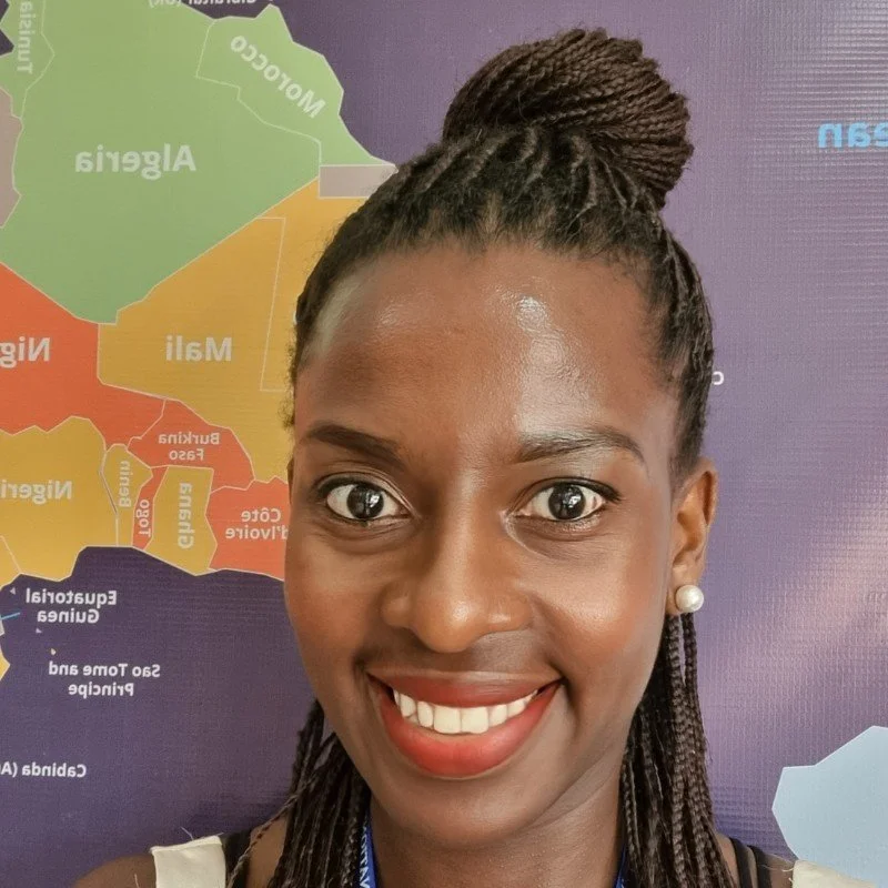Close-up of a smiling woman with braided hair in an updo, wearing pearl earrings, standing in front of a colorful map of Africa.