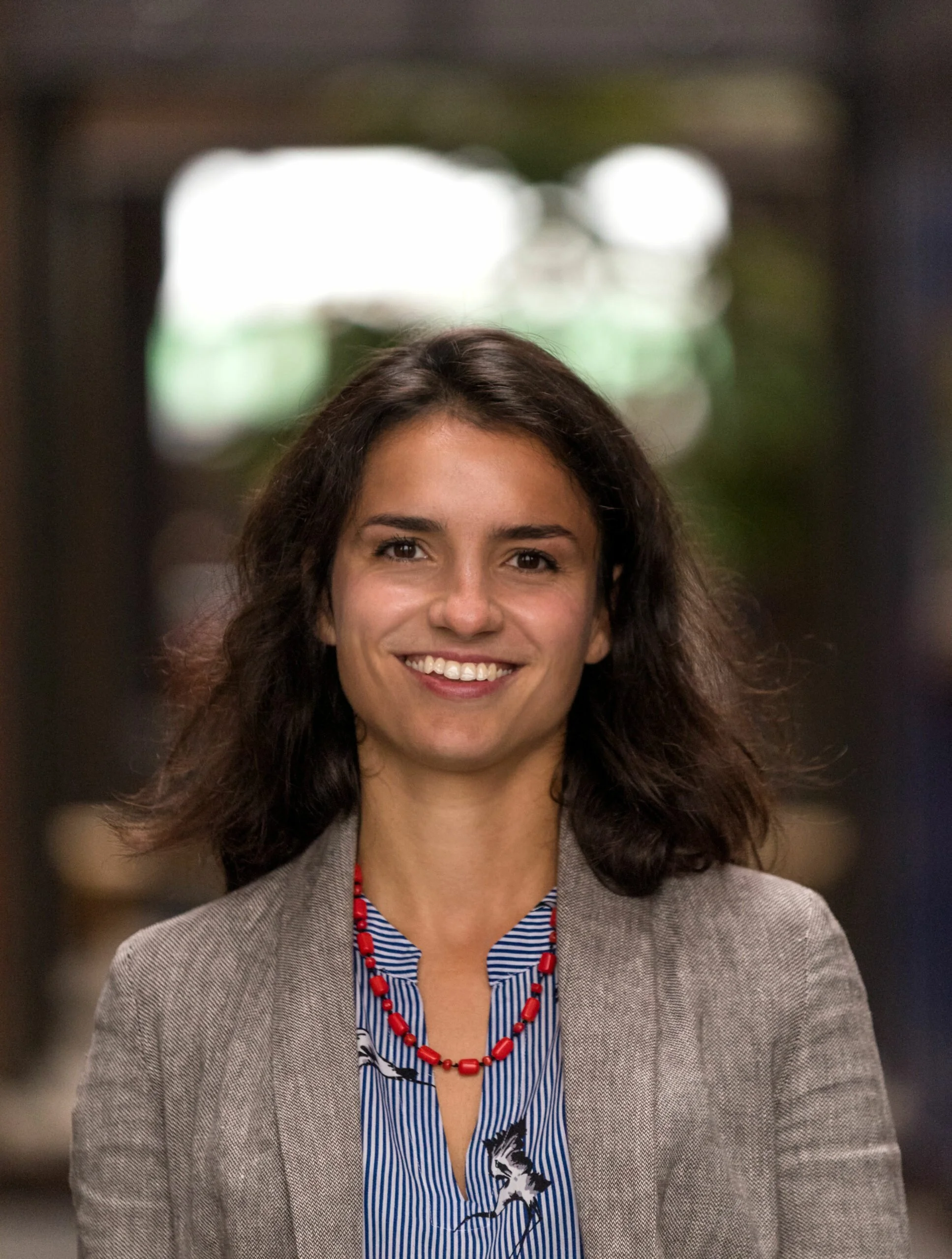 A woman with shoulder-length dark hair smiling at the camera, wearing a gray blazer, a blue striped shirt with a dog print, and a red beaded necklace, standing indoors with a blurred background.