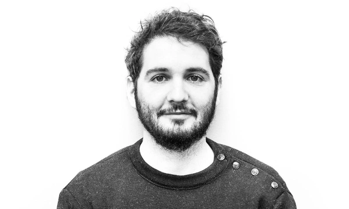 Black and white portrait of a young man with short, curly hair and a beard, wearing a dark sweater with decorative buttons on the shoulder.