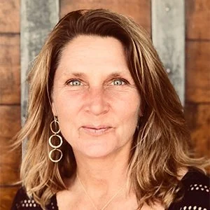 A woman with shoulder-length brown hair, wearing earrings, looking at the camera, with a wooden background.
