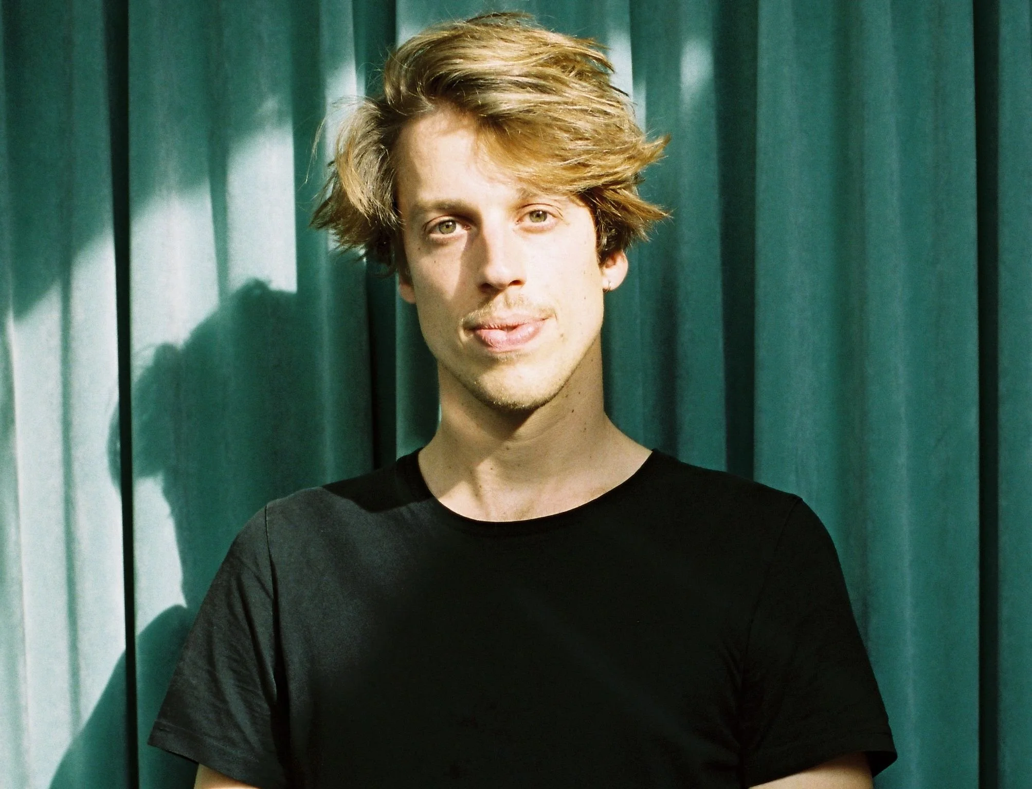 A young man with light skin, tousled blond hair, and a slight smile stands in front of teal curtains, wearing a black t-shirt.