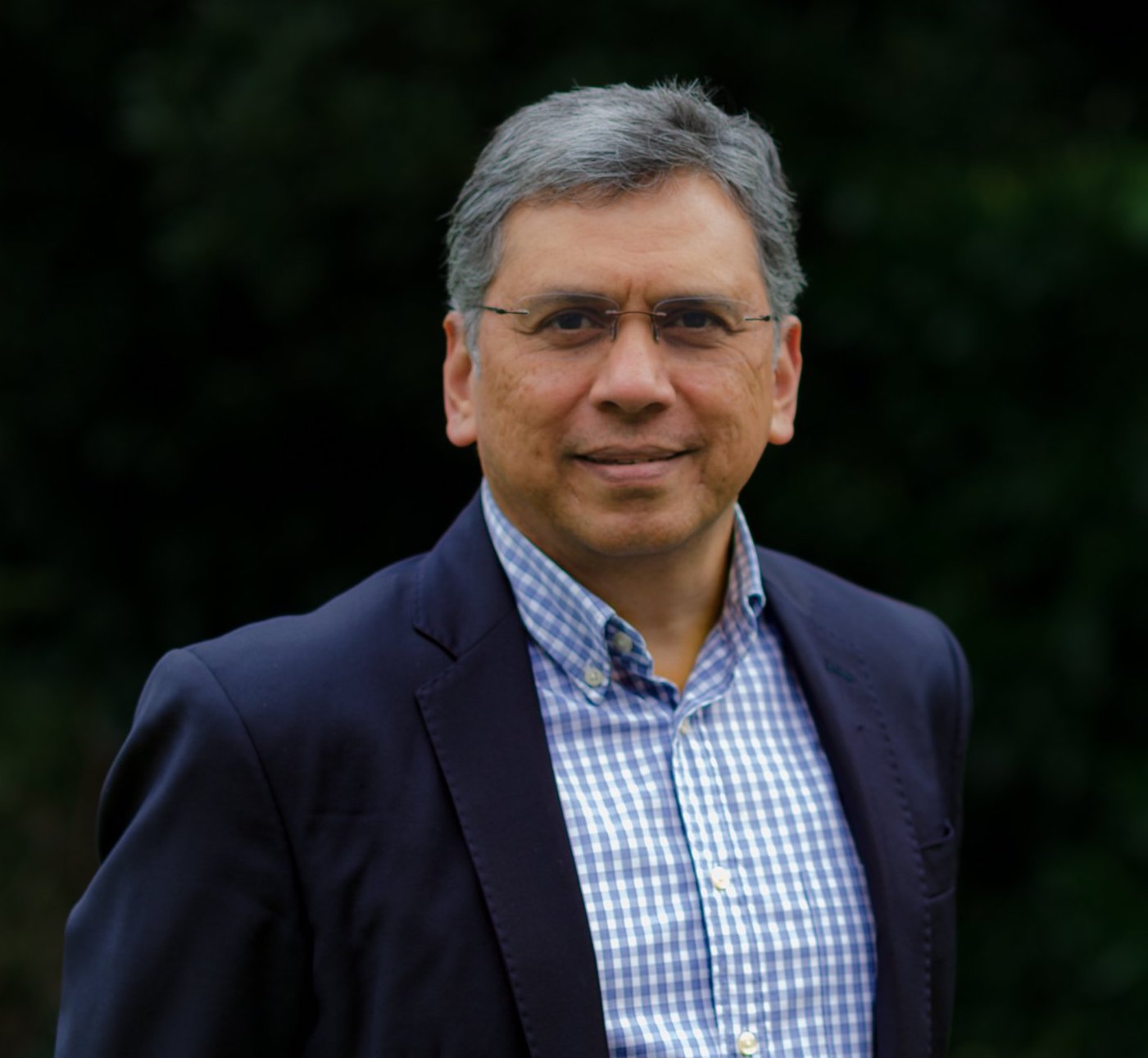 A middle-aged man with gray hair and glasses, wearing a navy blazer and a blue checkered shirt, standing outdoors against a dark green background.
