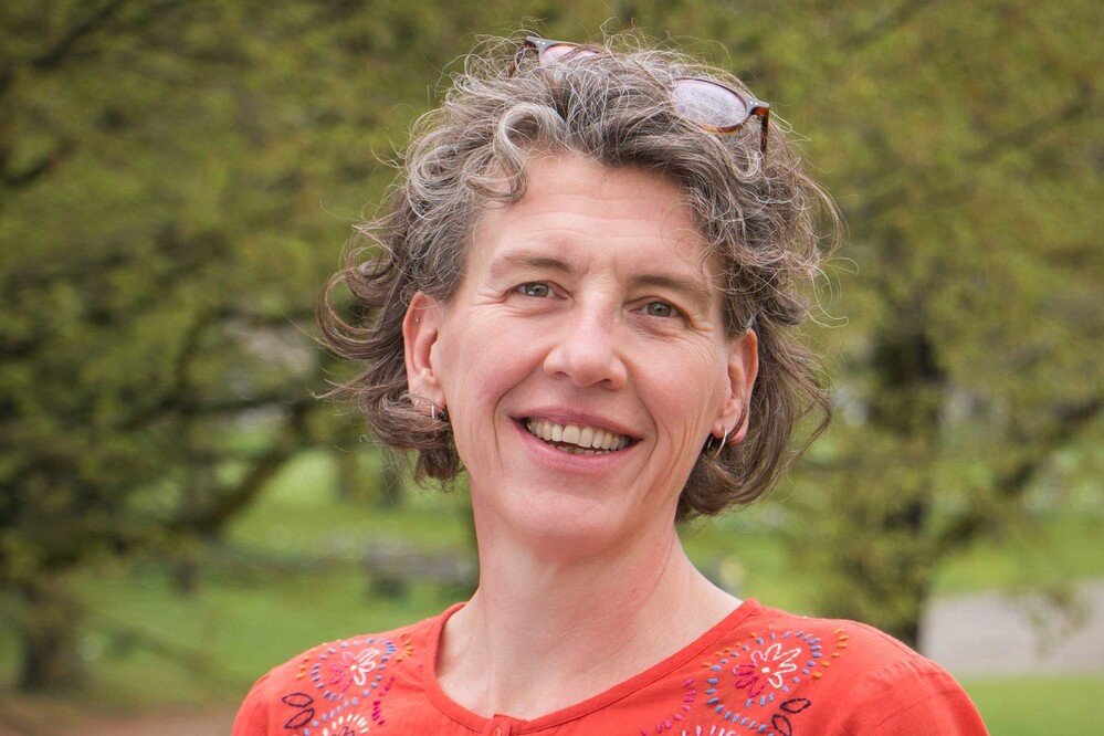 A woman with short, curly gray hair and glasses resting on her head, smiling outdoors in front of green trees, wearing an embroidered orange top.
