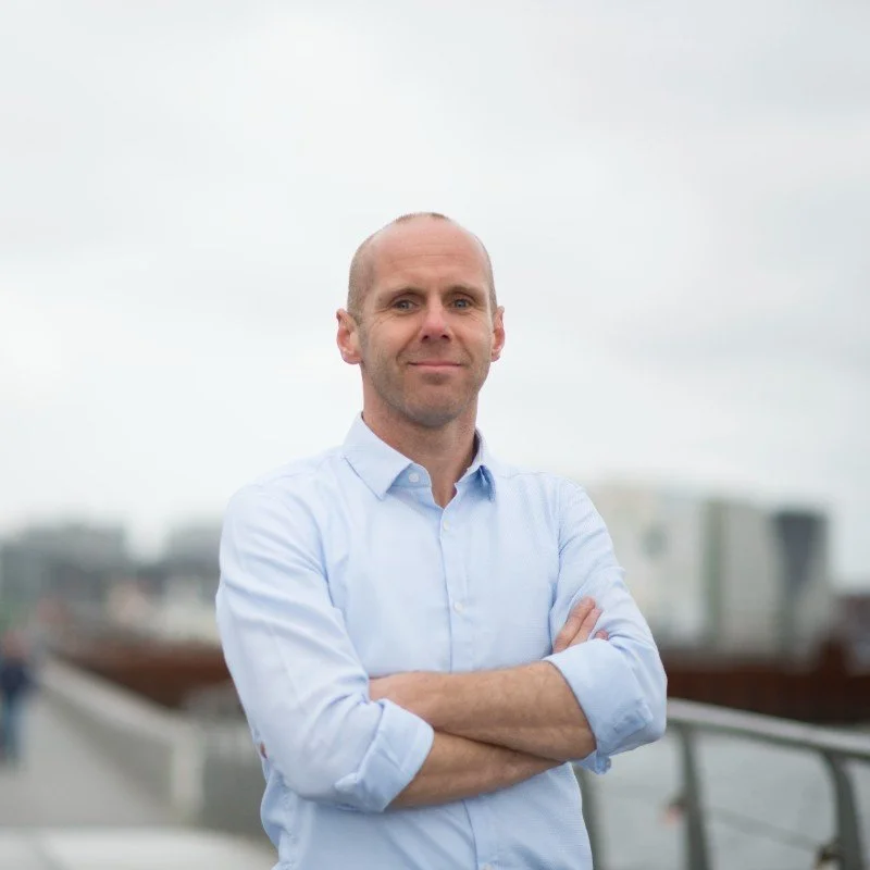 A man with a shaved head and a light blue button-up shirt standing outdoors with arms crossed, smiling slightly with city buildings in the background.