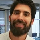 A smiling man with dark hair and a beard, indoors with large windows in the background.