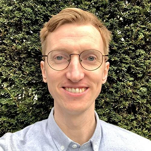 A young man with light skin, blond hair, and glasses, smiling outdoors in front of a bush.