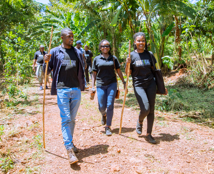 Group of young people walking through a lush, green jungle trail while carrying sticks and wearing matching black t-shirts with the word 'ROOTICAL' on them.