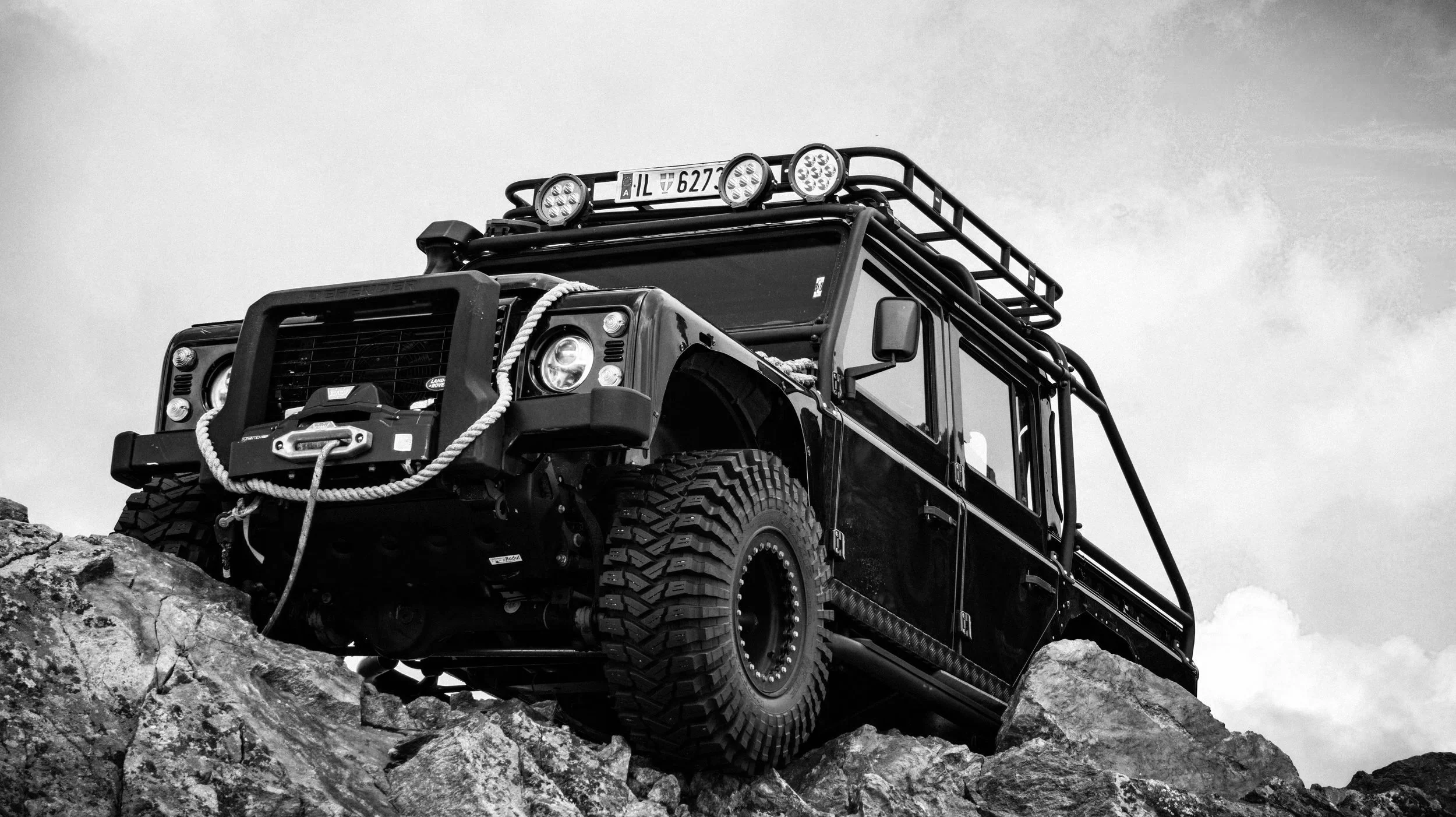 A black off-road vehicle with rugged tires, bumper, and roof rack, positioned on rocky terrain under cloudy sky.