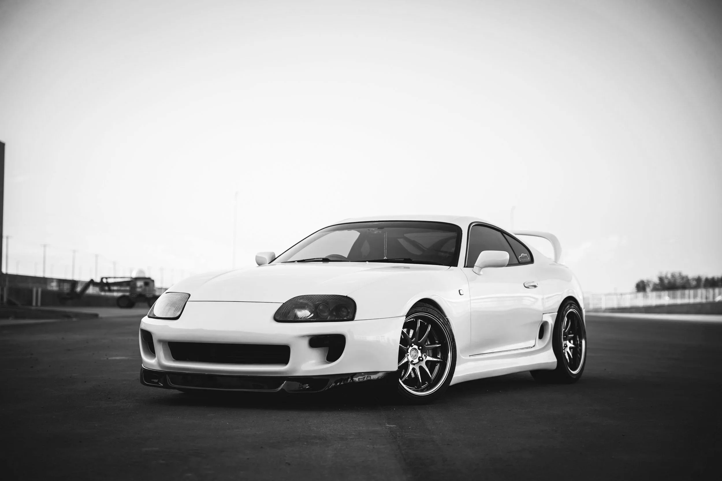 Black and white photo of a white sports car with sleek design, large rear wing, and aftermarket wheels parked on a racetrack.