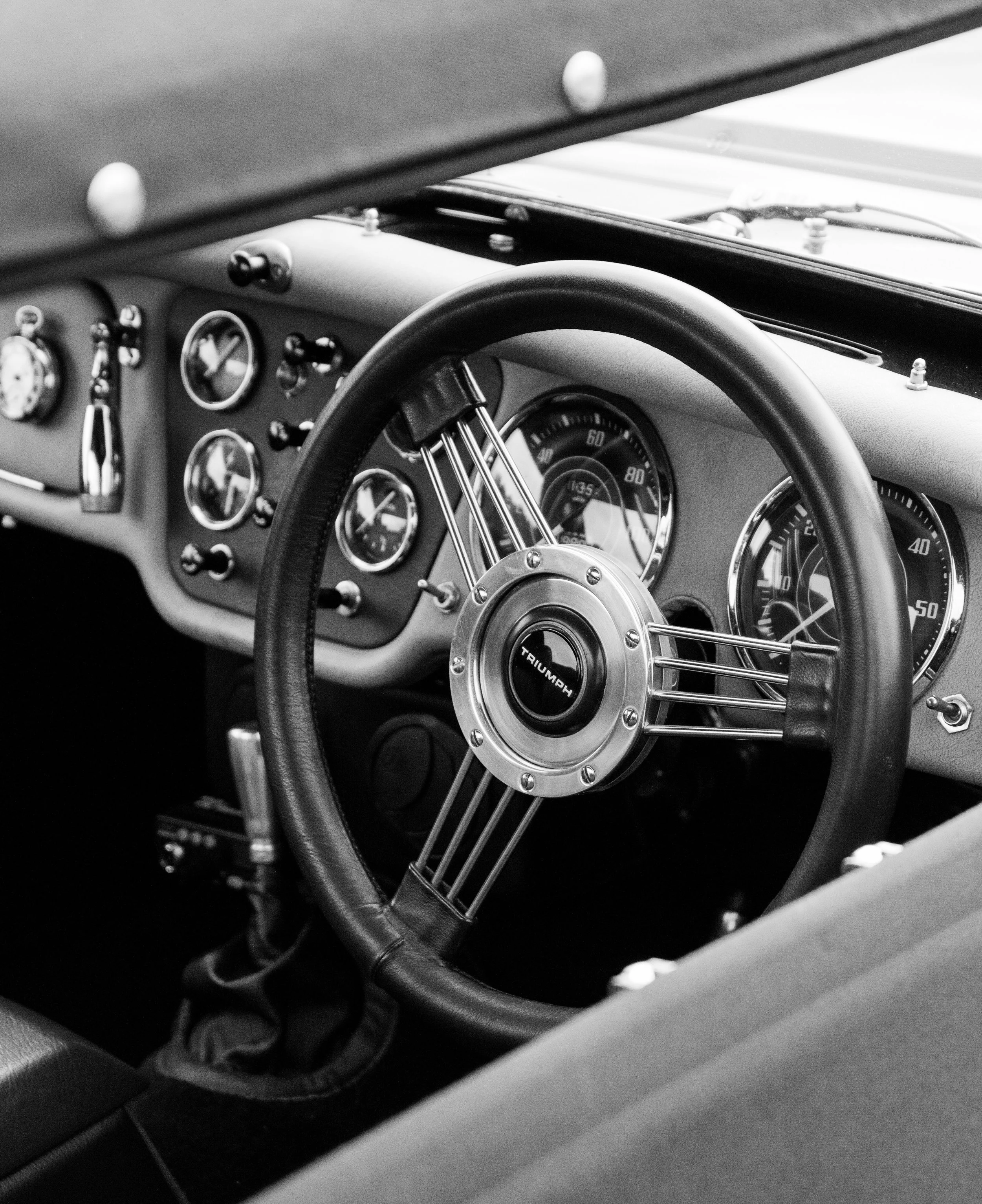 Interior of a vintage car, showing the dashboard with analog gauges, a steering wheel with a Triumph logo, and various controls in black and white.