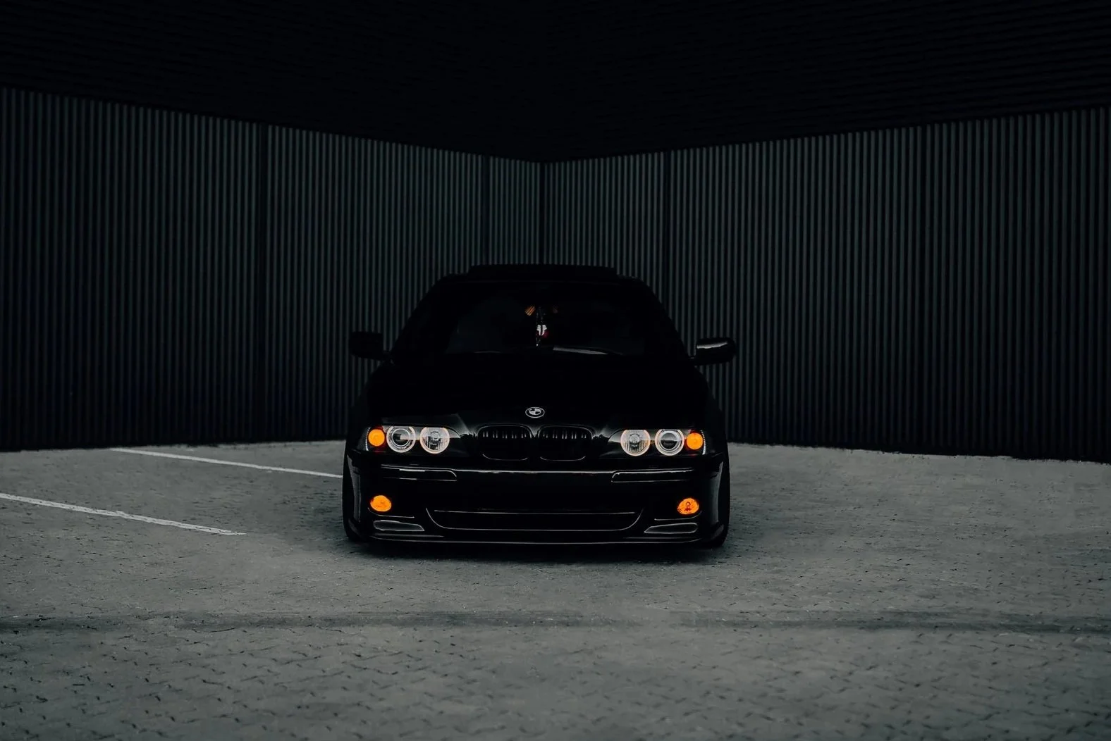 Black sports car parked in a dark, covered parking lot with corrugated metal walls.