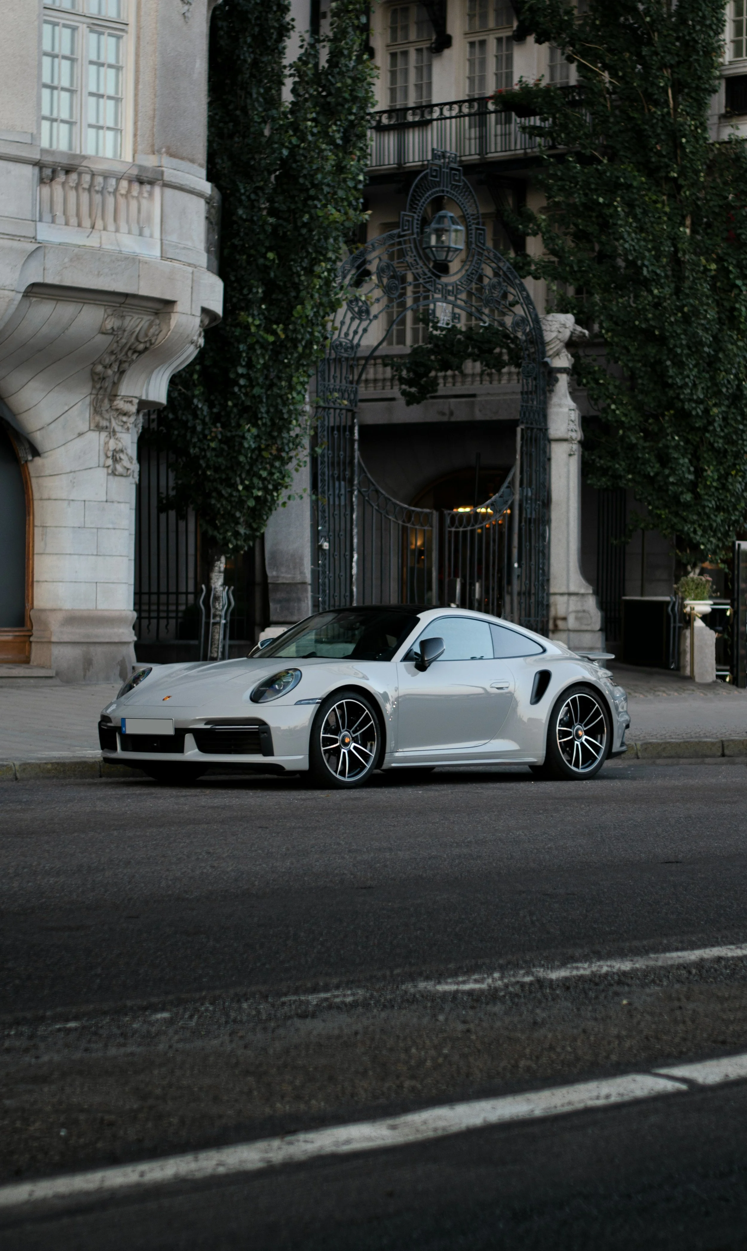 A silver Porsche 911 sports car is parked on the street in front of a historic building with an ornate iron gate and ivy-covered columns.