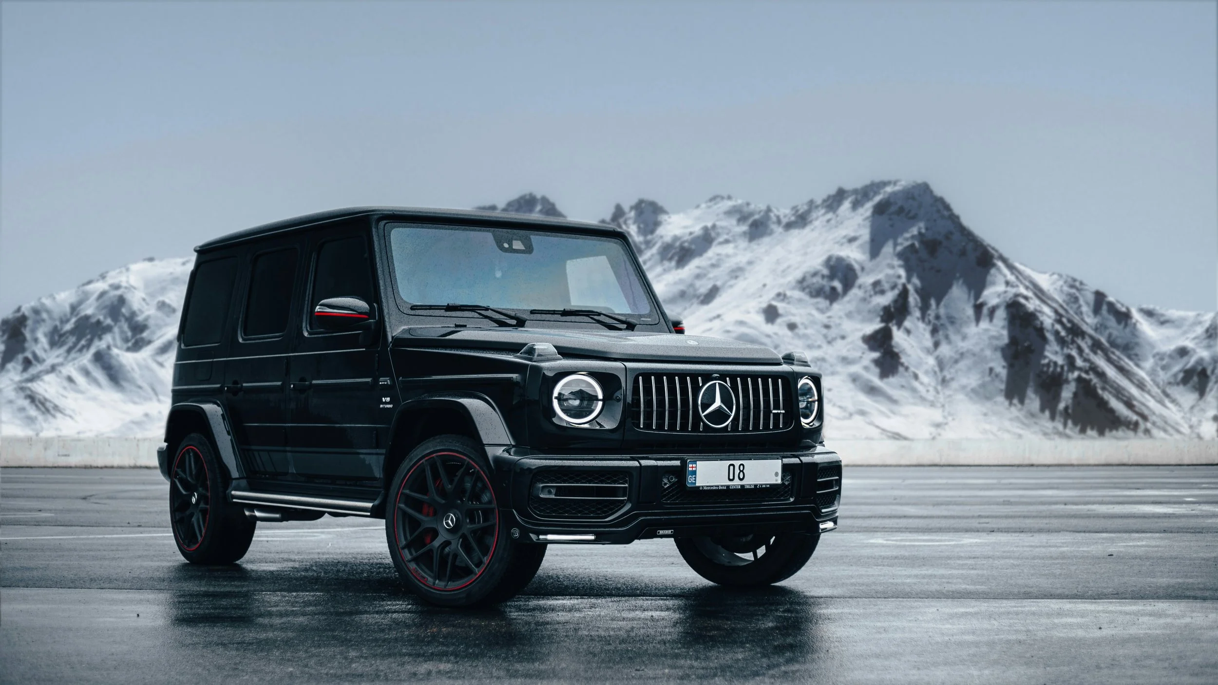 Black Mercedes-Benz G-Class SUV parked on a wet asphalt surface with snow-covered mountains in the background.