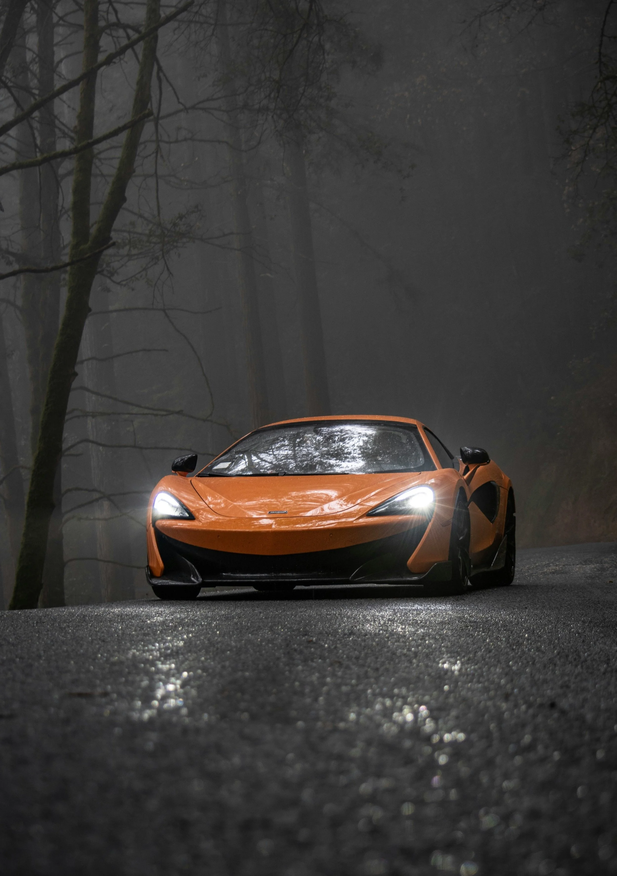An orange sports car with its headlights on, parked on a wet, dark forest road with fog and trees in the background.