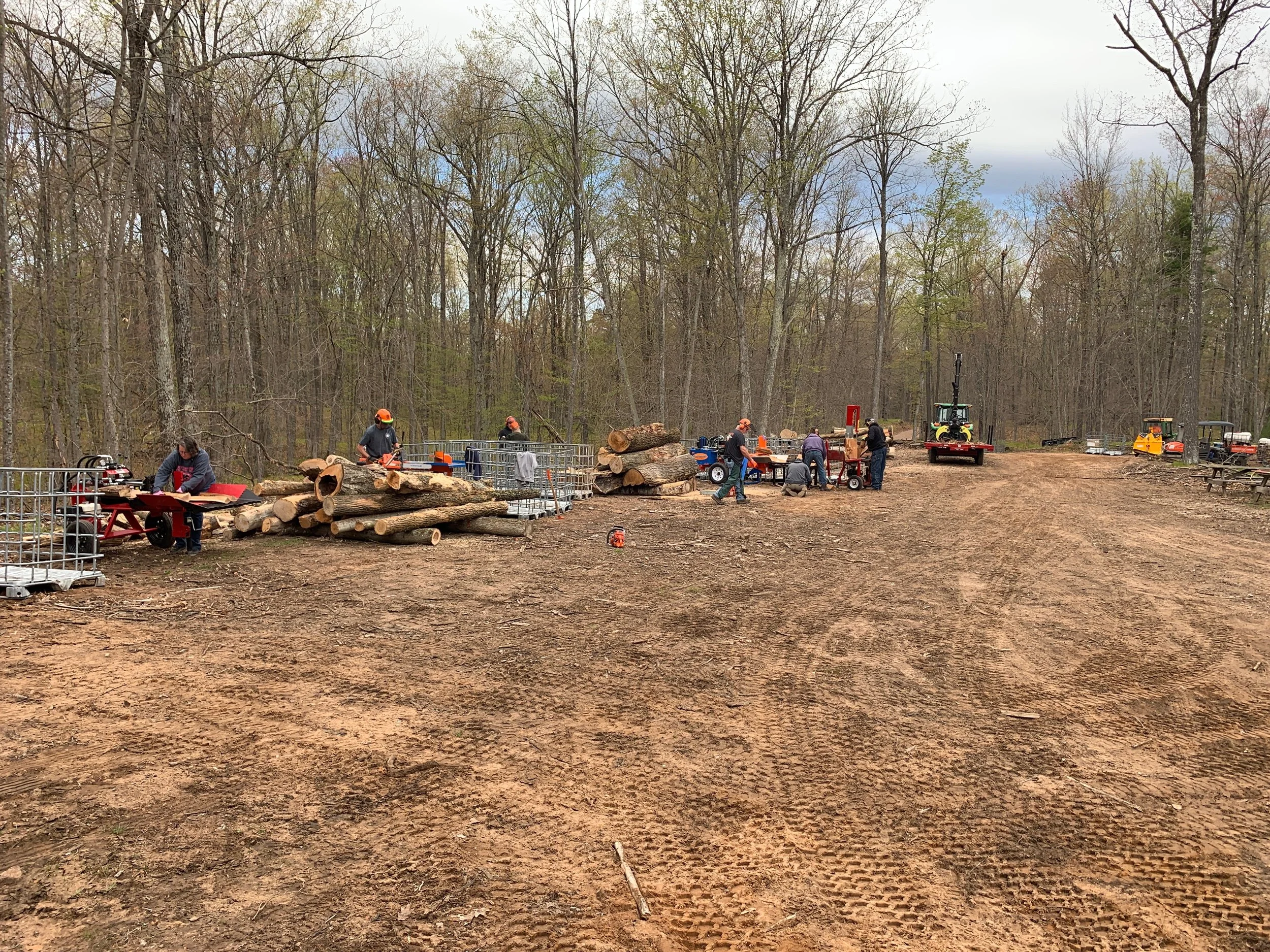 Group of people in a wooded outdoor area working on logging and wood processing tasks, with logs, equipment, and machinery visible, and trees in the background.