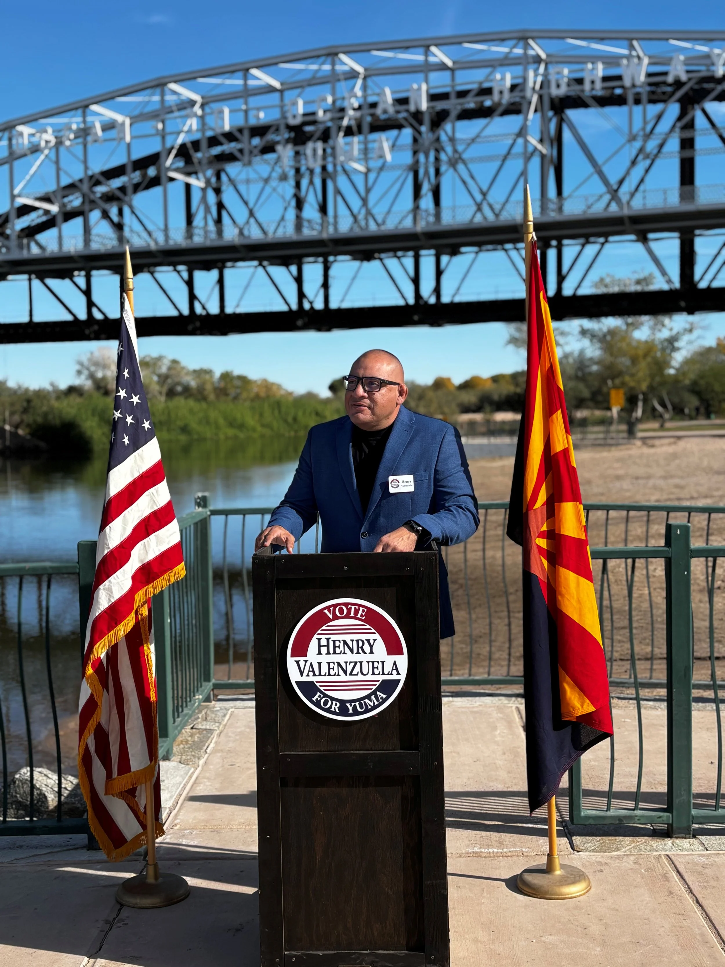 Man in blue suit at podium with campaign sign that reads 'Vote Henry Valenzuela for Yuma', flanked by American and Arizona flags, outdoors near a river and bridge.