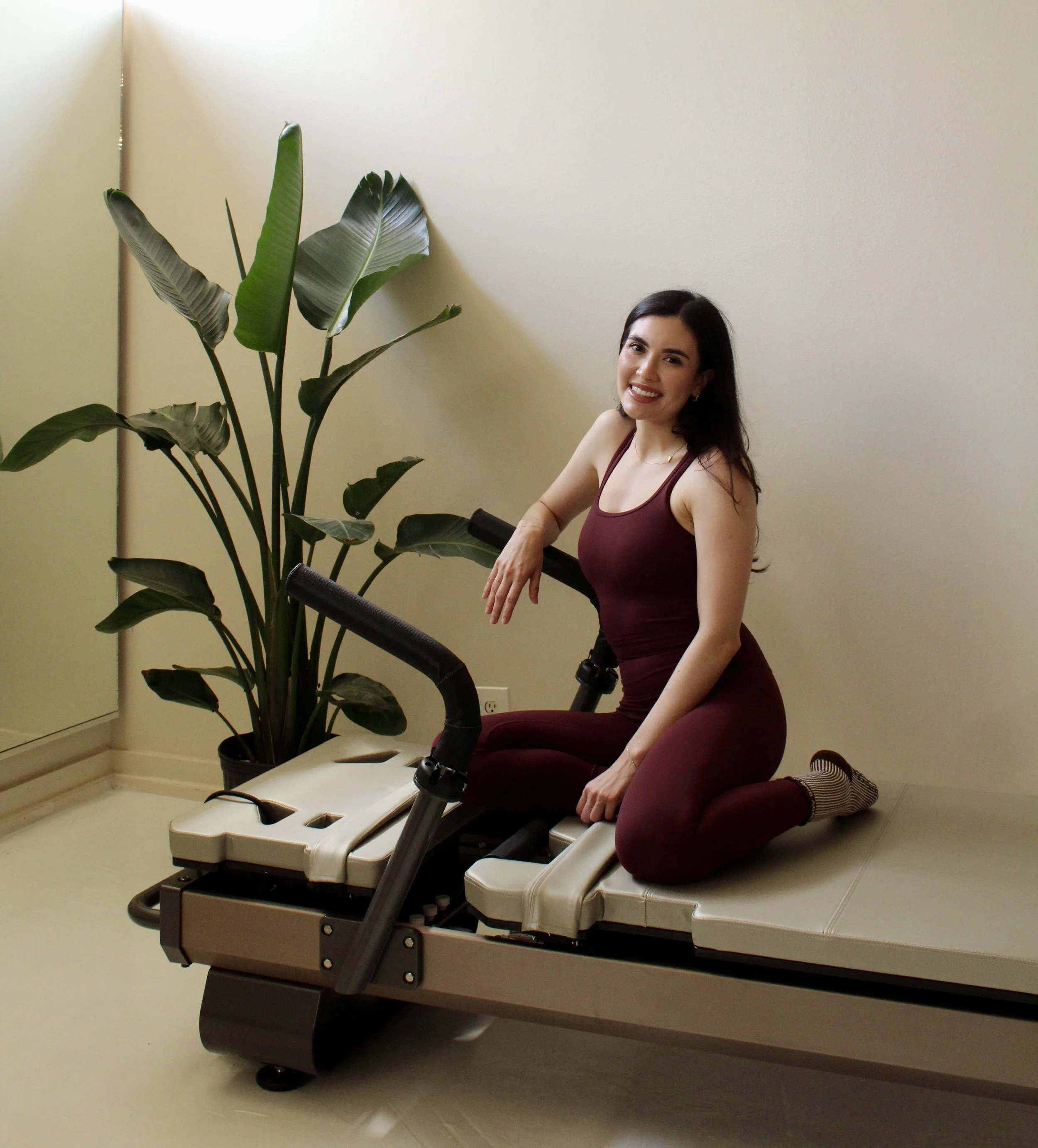 Pilates instructor seated on a reformer machine, smiling, wearing a maroon athletic outfit, with a tropical plant in the background