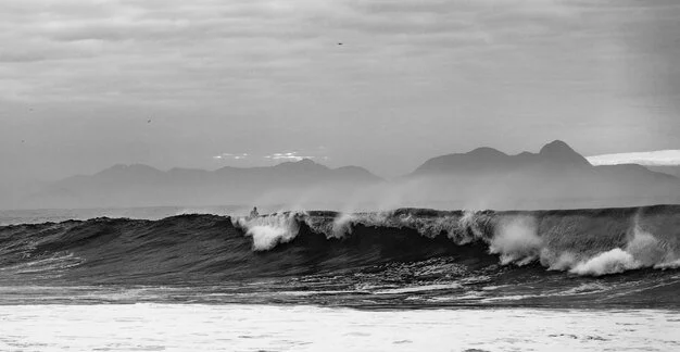 Waves crashing on the ocean with mountains in the distance under a cloudy sky.