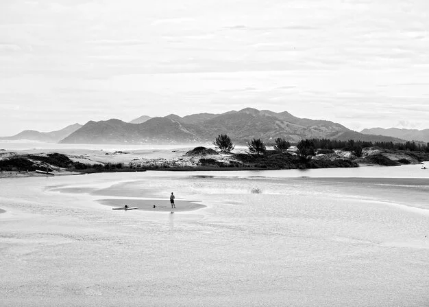 A person stands on a sandbar in a beach with mountains in the background, in black and white.
