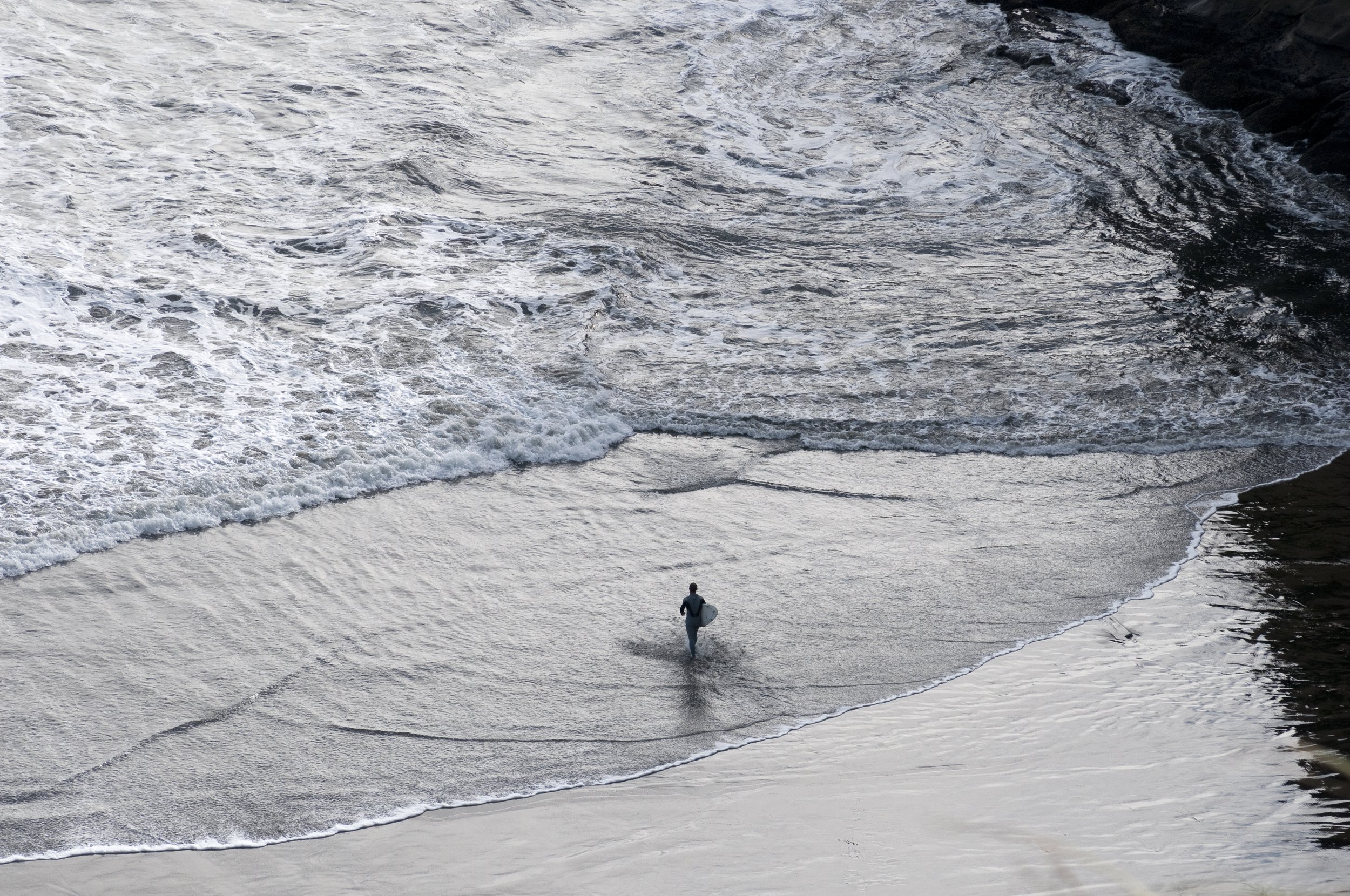 A person holding a surfboard walking along a beach shoreline with waves crashing nearby.