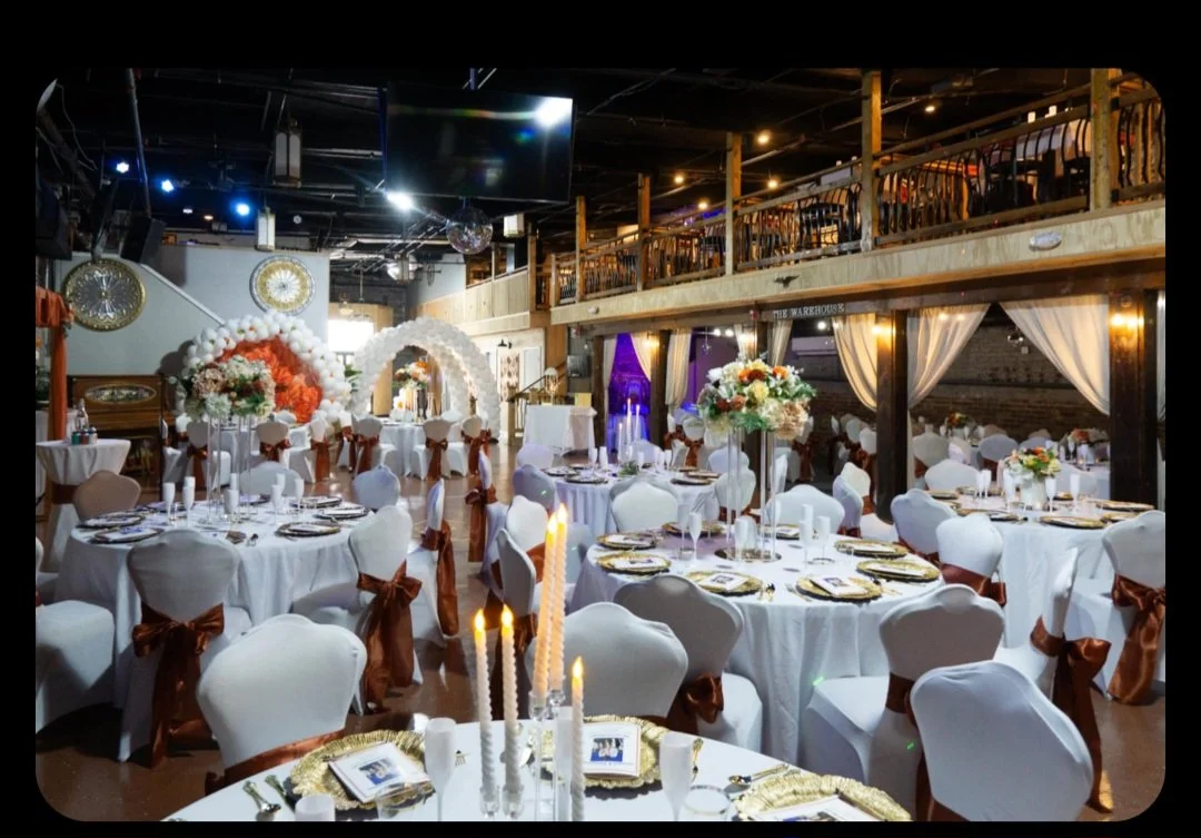 Wedding reception setup with round tables decorated with white tablecloths, floral centerpieces, and chairs with white covers and brown sashes. An arch made of white balloons and flower arrangements is in the background. The venue has a second-floor balcony and warm lighting.