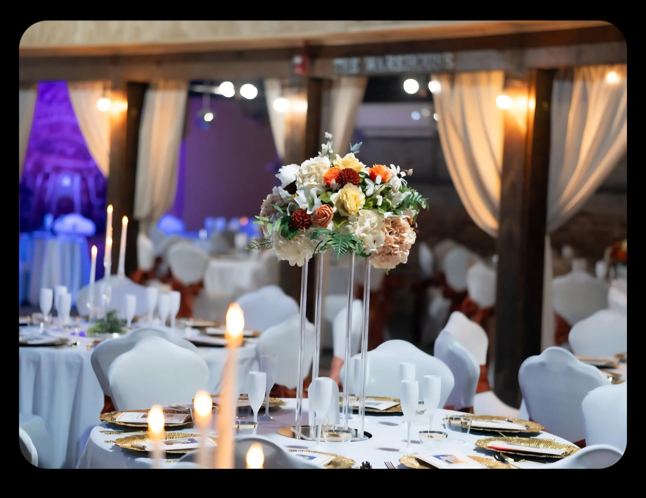 Elegant banquet hall decorated for a formal event with round tables draped in white tablecloths, set with gold chargers, champagne glasses, and place settings, featuring a tall floral centerpiece of white, yellow, orange, and red flowers, with soft lighting and draped curtains in the background.