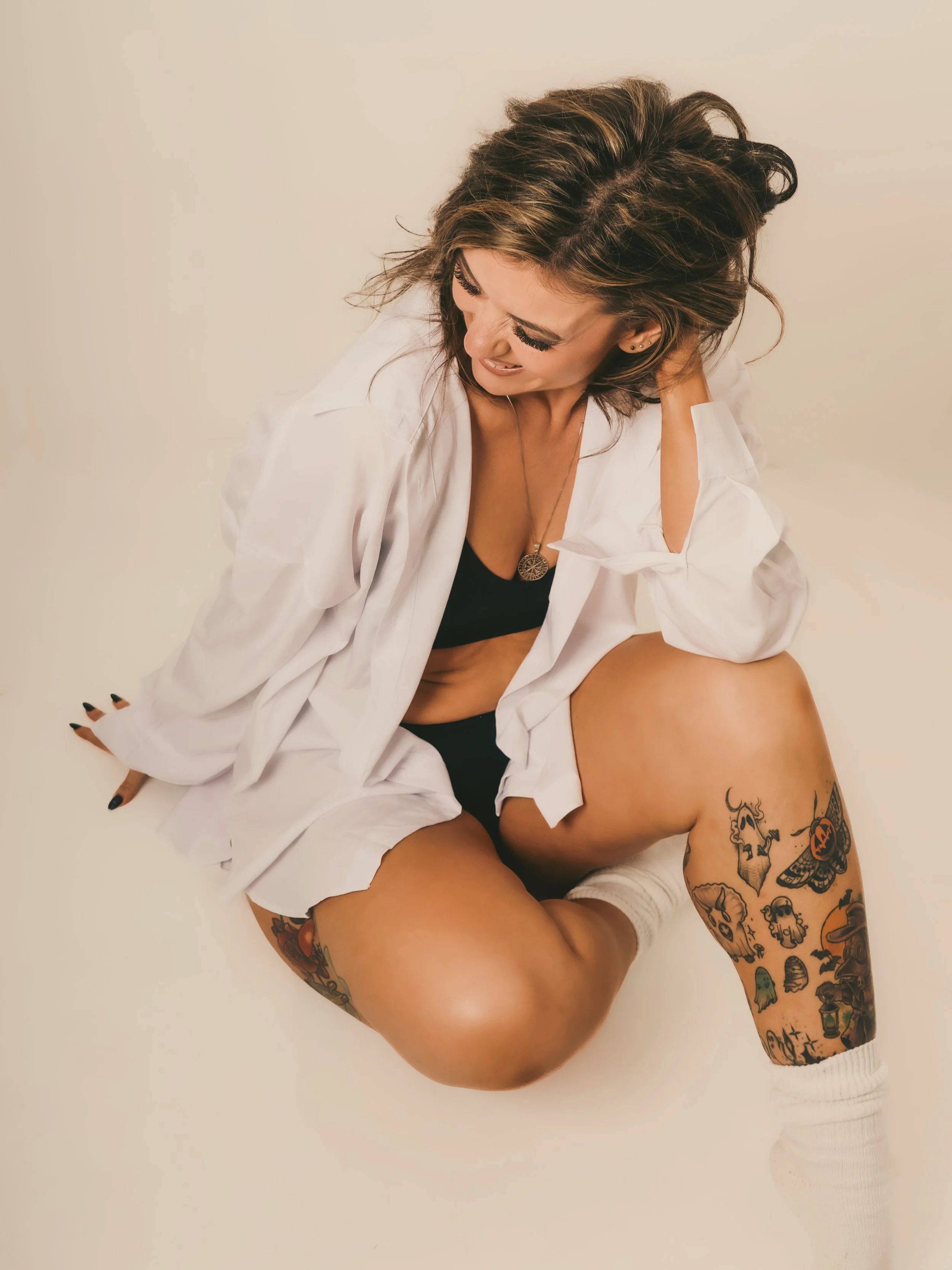 A woman sitting on the floor with her legs bent, wearing a white shirt, black bra, and white socks, smiling and looking down.