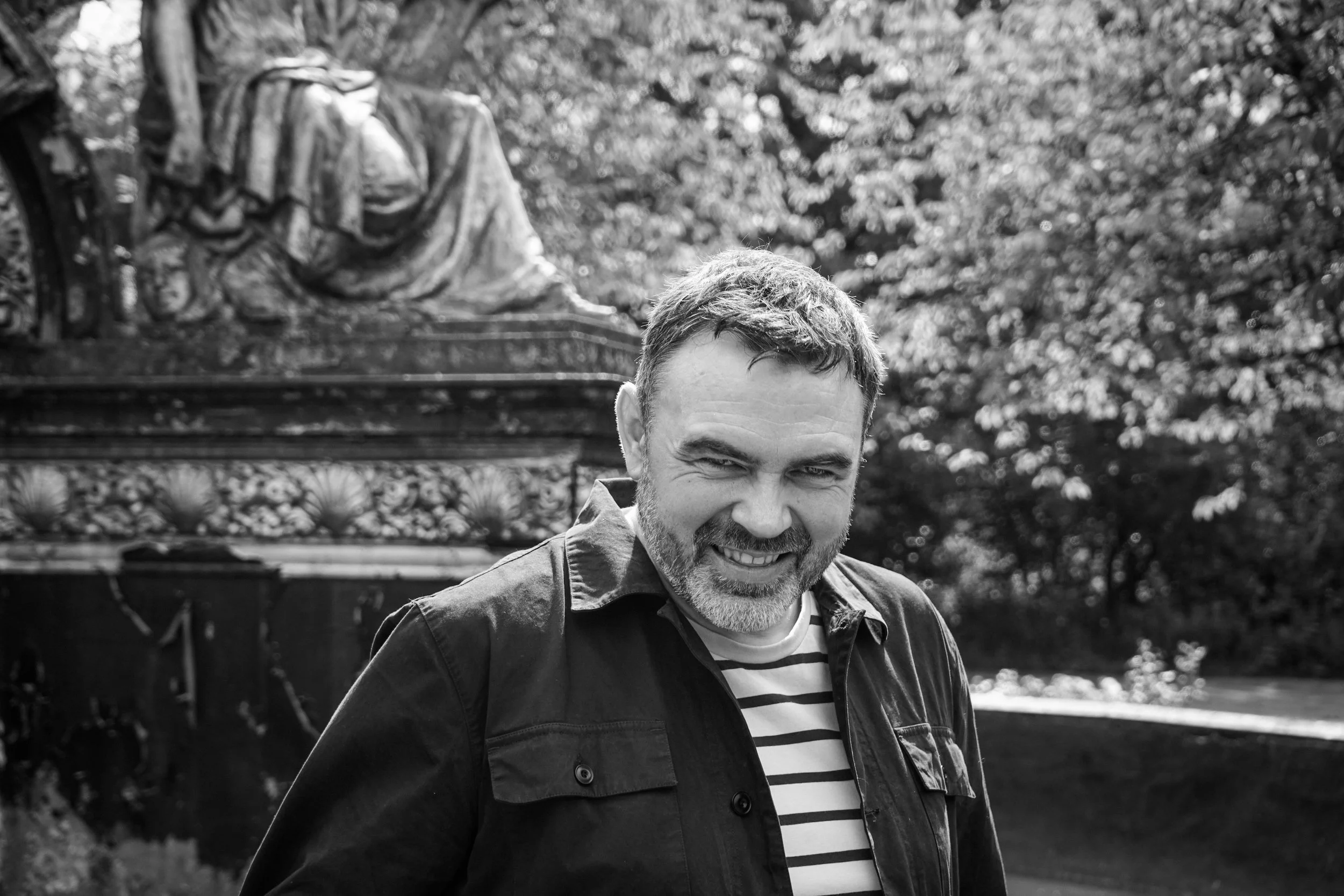 A man with a beard and short hair smiling outdoors in front of a sculpture of a seated figure, with trees in the background.