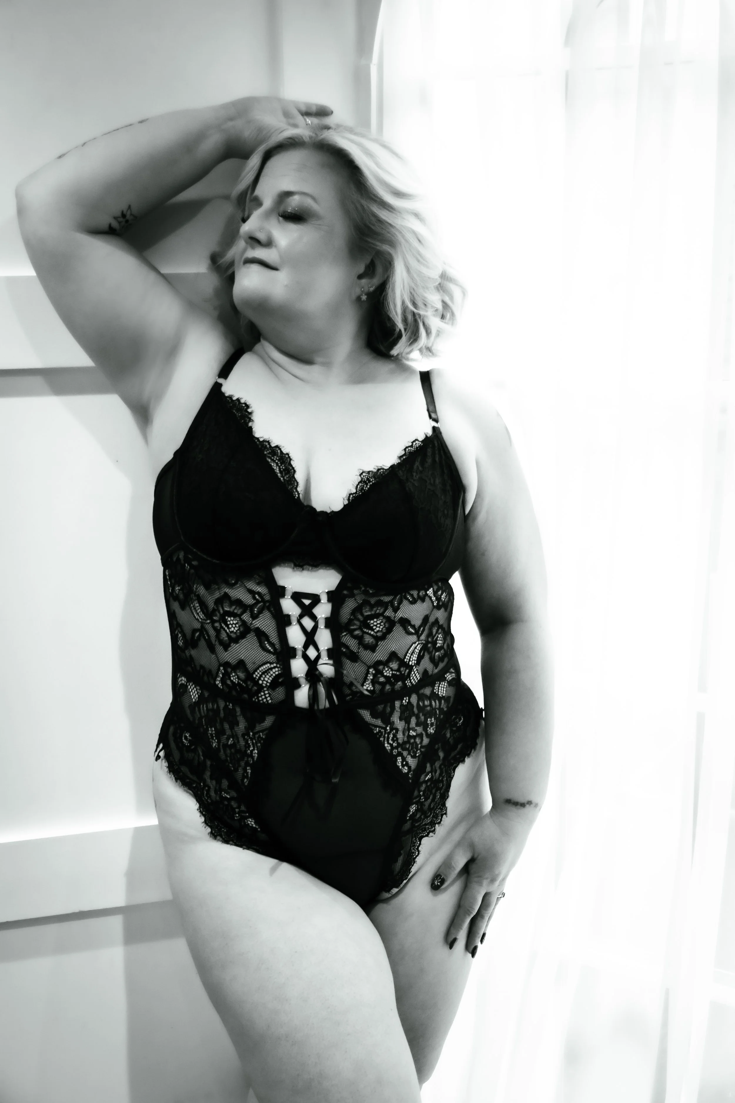 A woman in black lace lingerie striking a pose with one arm raised and resting on her head, standing in front of a light-colored background and sheer curtains.