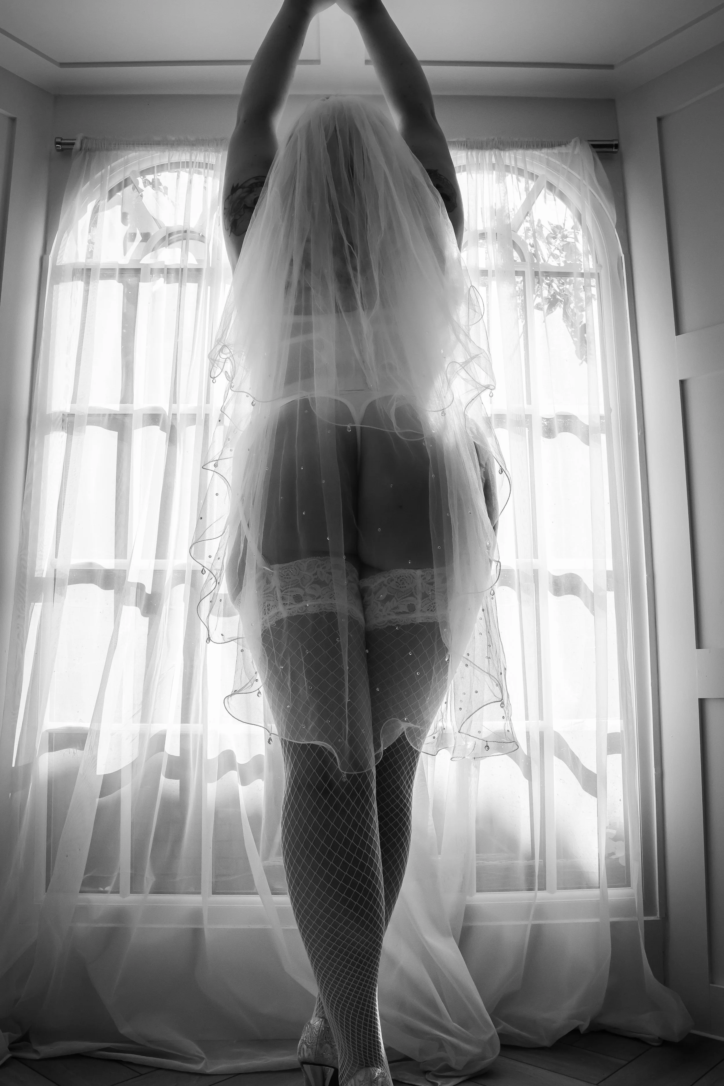 A woman in wedding lingerie, fishnet stockings, and high heels standing behind a sheer curtain near a window, with her arms raised above her head.