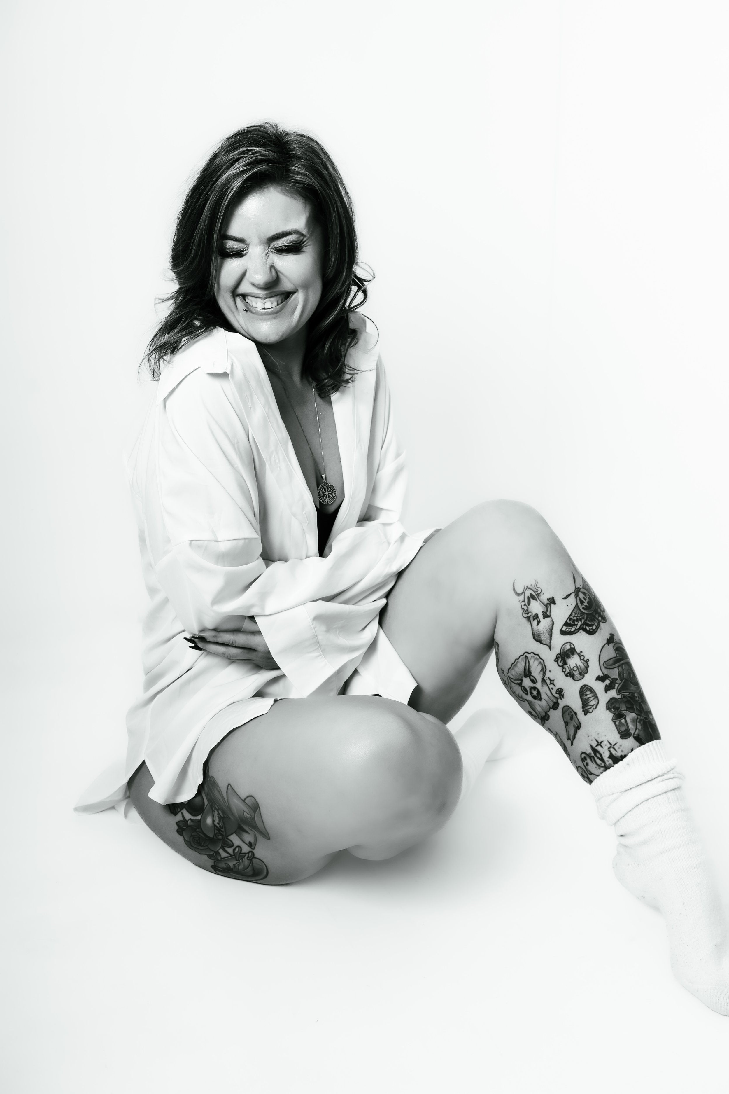 Black and white photo of a woman sitting on the floor, smiling with eyes closed, wearing a white shirt and tattoos on her legs, also wearing a white sock on one foot.