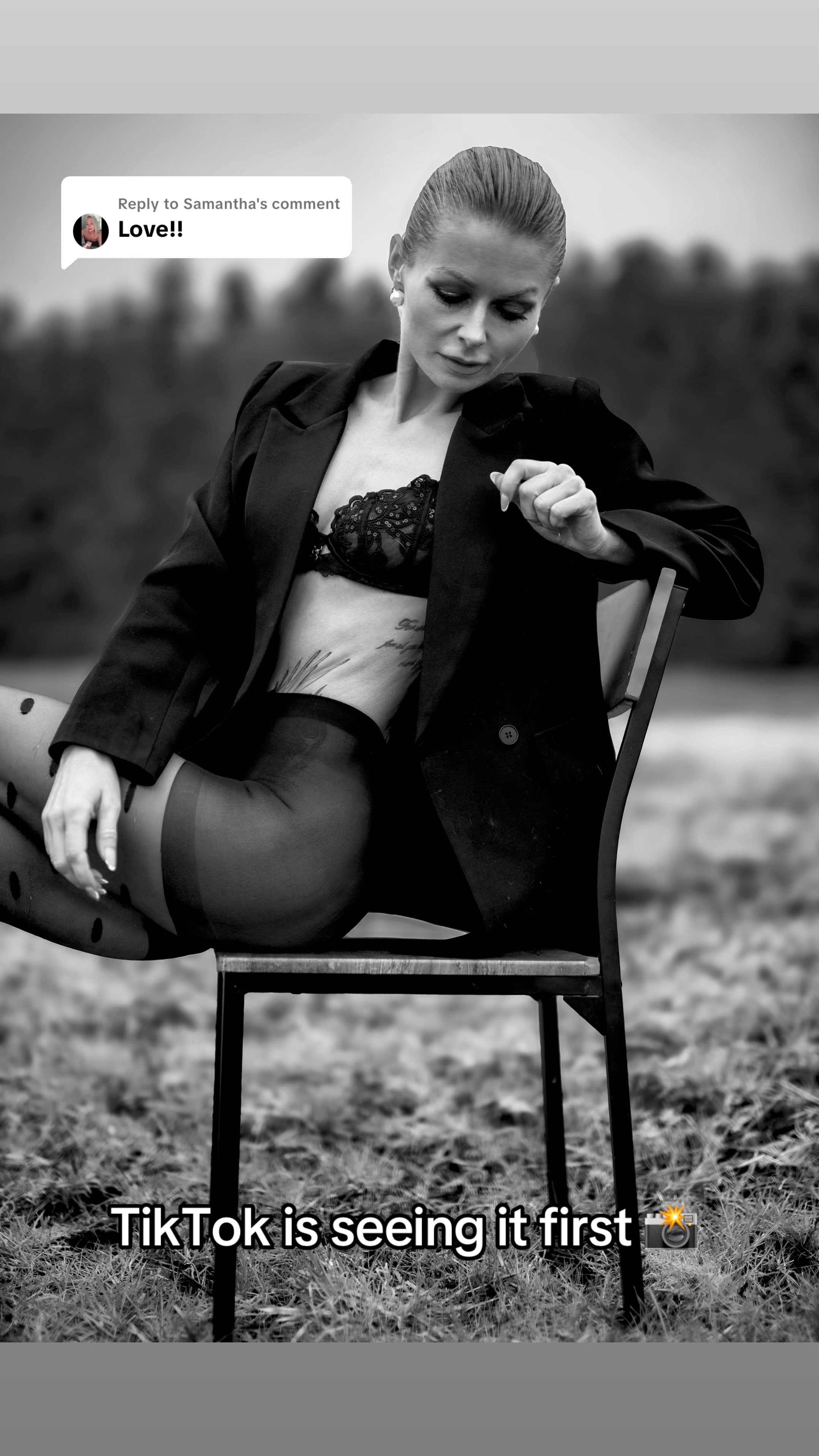 A black-and-white photo of a woman sitting on a chair outdoors, wearing a black blazer, lace bra, and stockings with watermelon seed patterns. She has her hair tied back and is looking down. A speech bubble says, 'Love!!' in response to a comment. Te