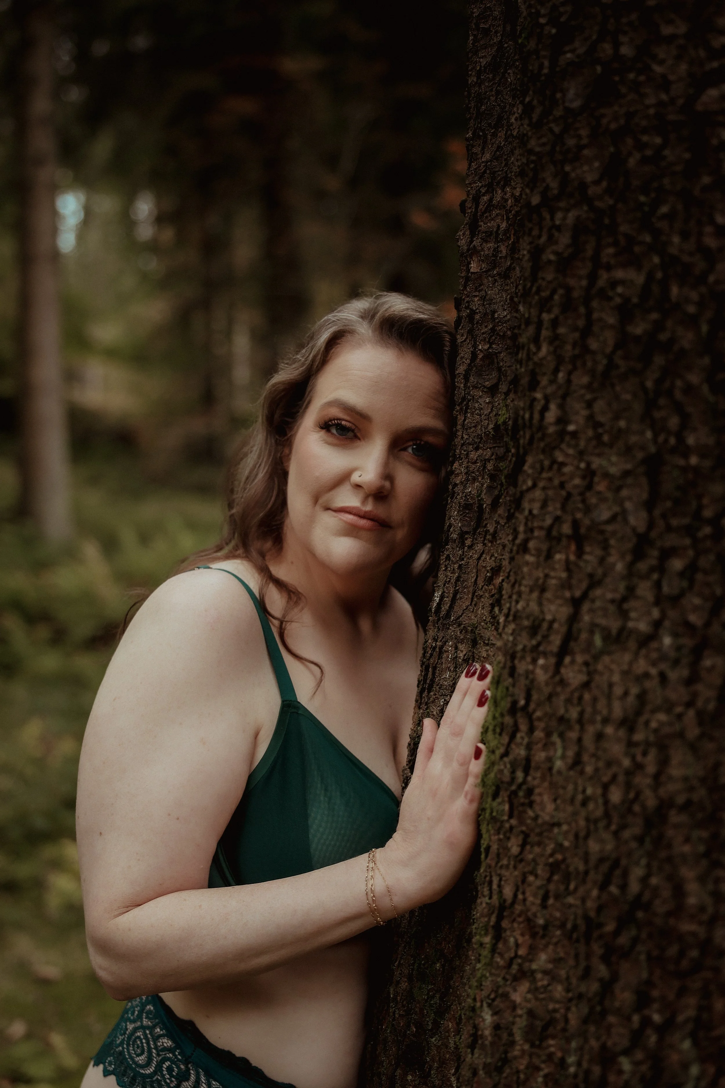 A woman with wavy brown hair, wearing a dark green and black lingerie, stands outdoors in a forest, leaning against a tree and looking at the camera.