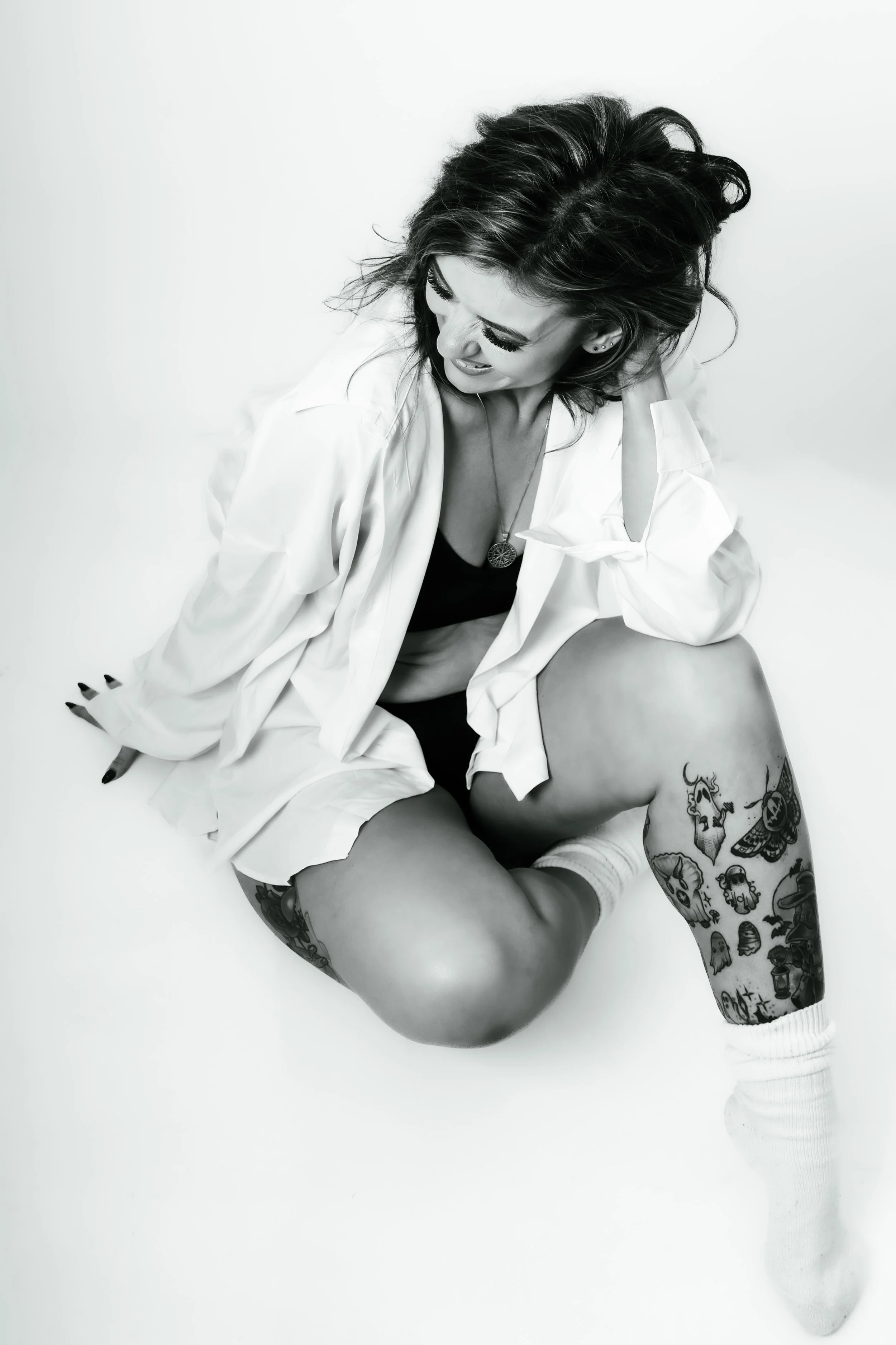 Black and white photo of a woman with tattoos sitting on the floor, wearing a white shirt, black crop top, and white socks, with her head tilted and looking down.