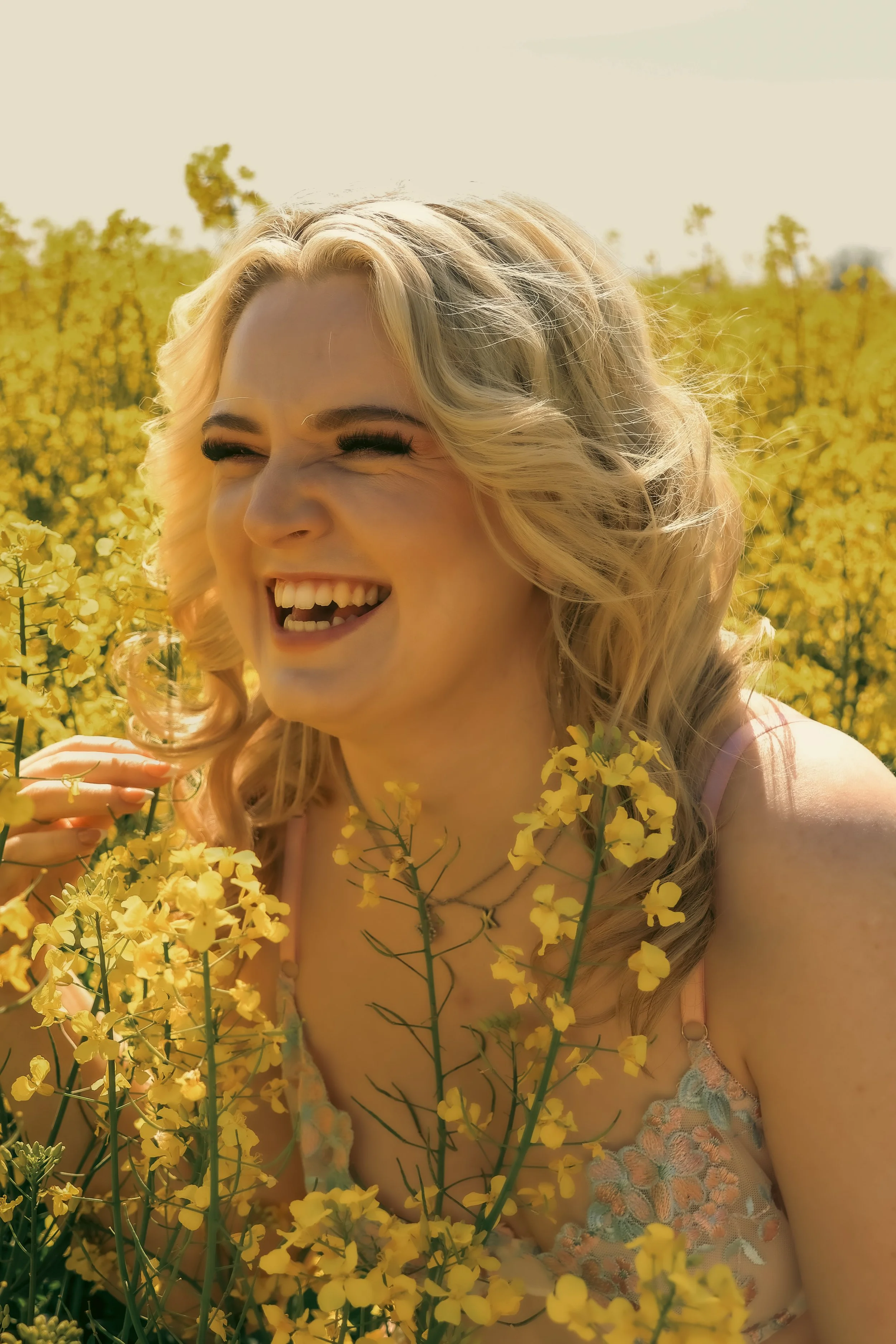 A woman with blonde curly hair smiling joyfully among yellow flowers in a field, wearing a light-colored top.