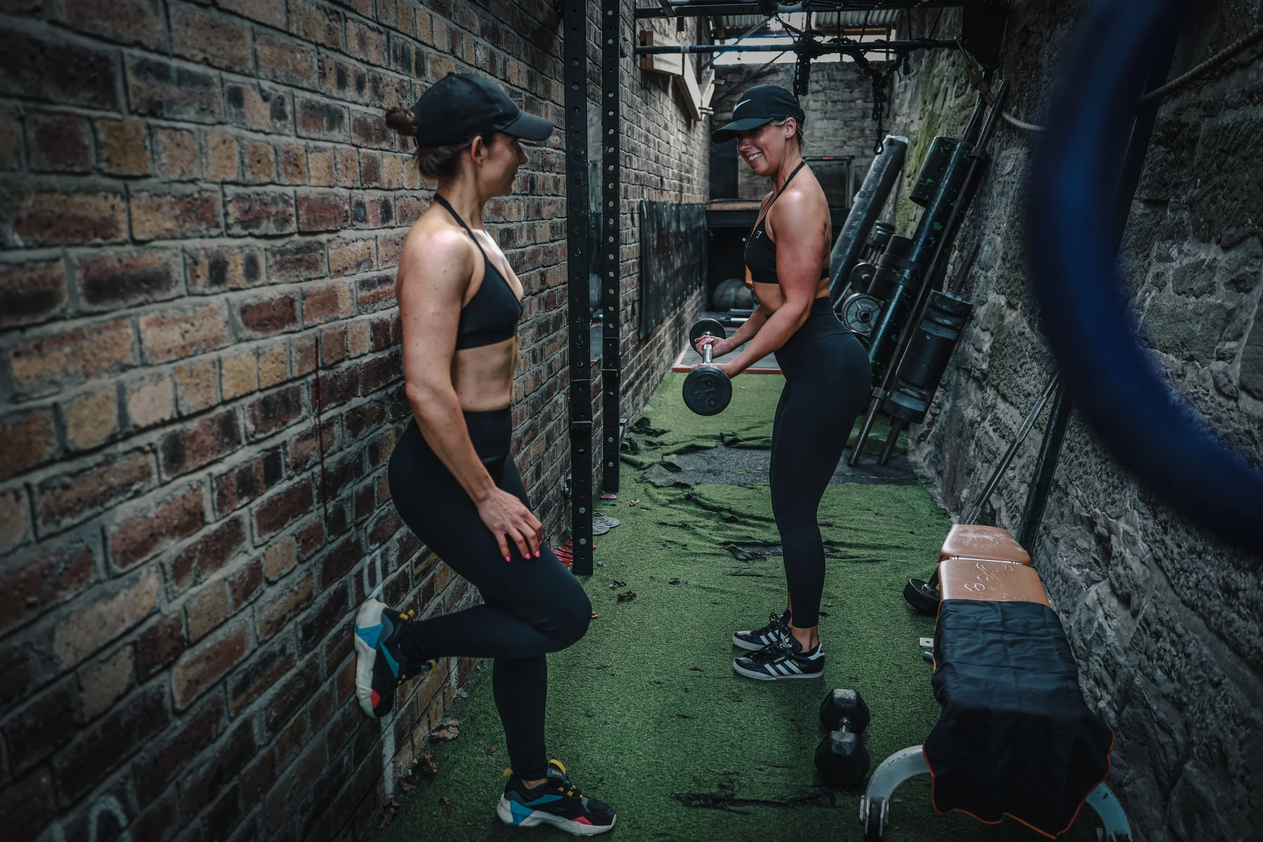 Two women in workout attire exercising in a narrow outdoor gym space with brick and stone walls, grass flooring, and workout equipment. One is holding dumbbells while the other is stretching.