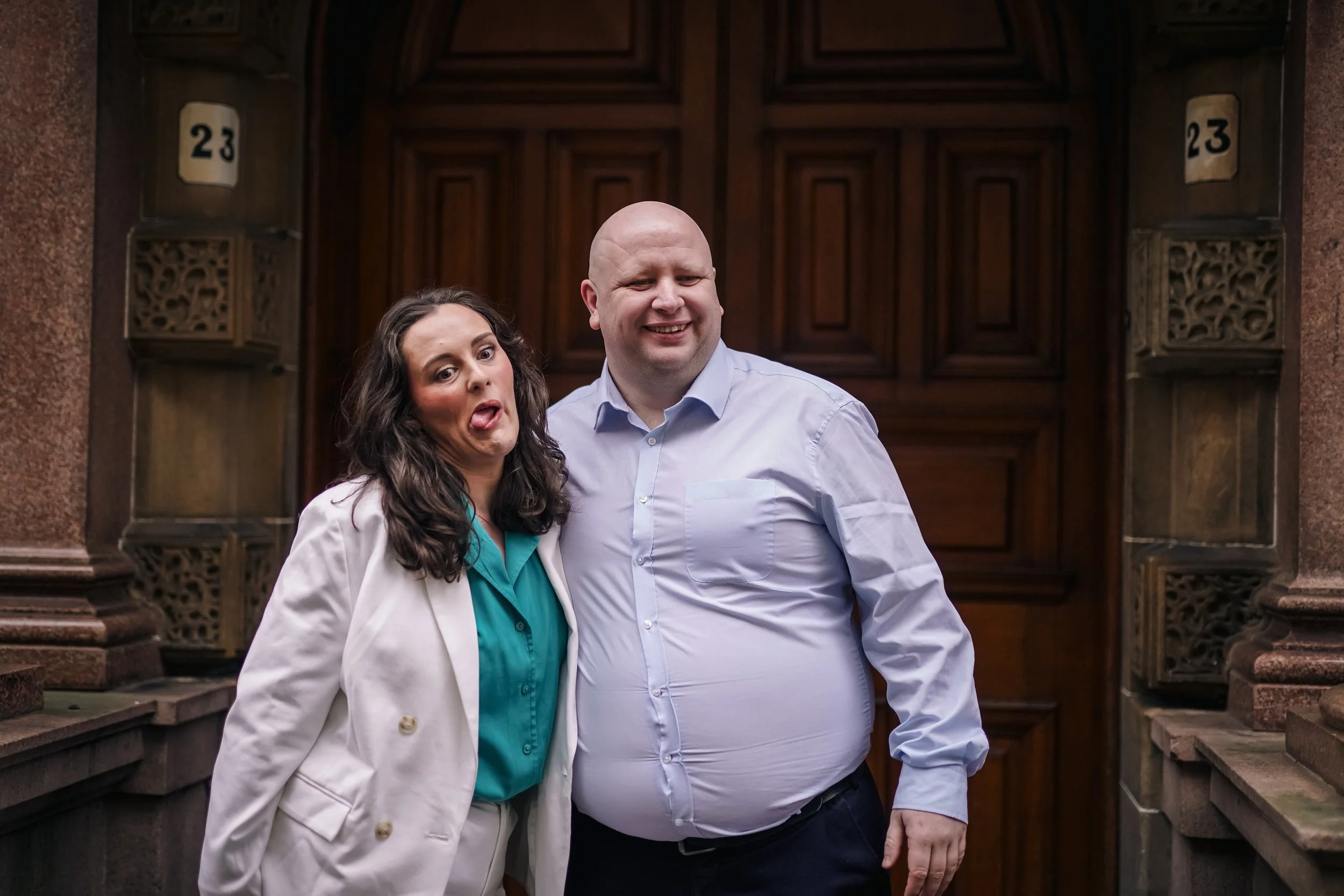 A woman making a funny face and sticking out her tongue next to a smiling bald man. They are standing in front of a large wooden door with the number 23 on both sides.