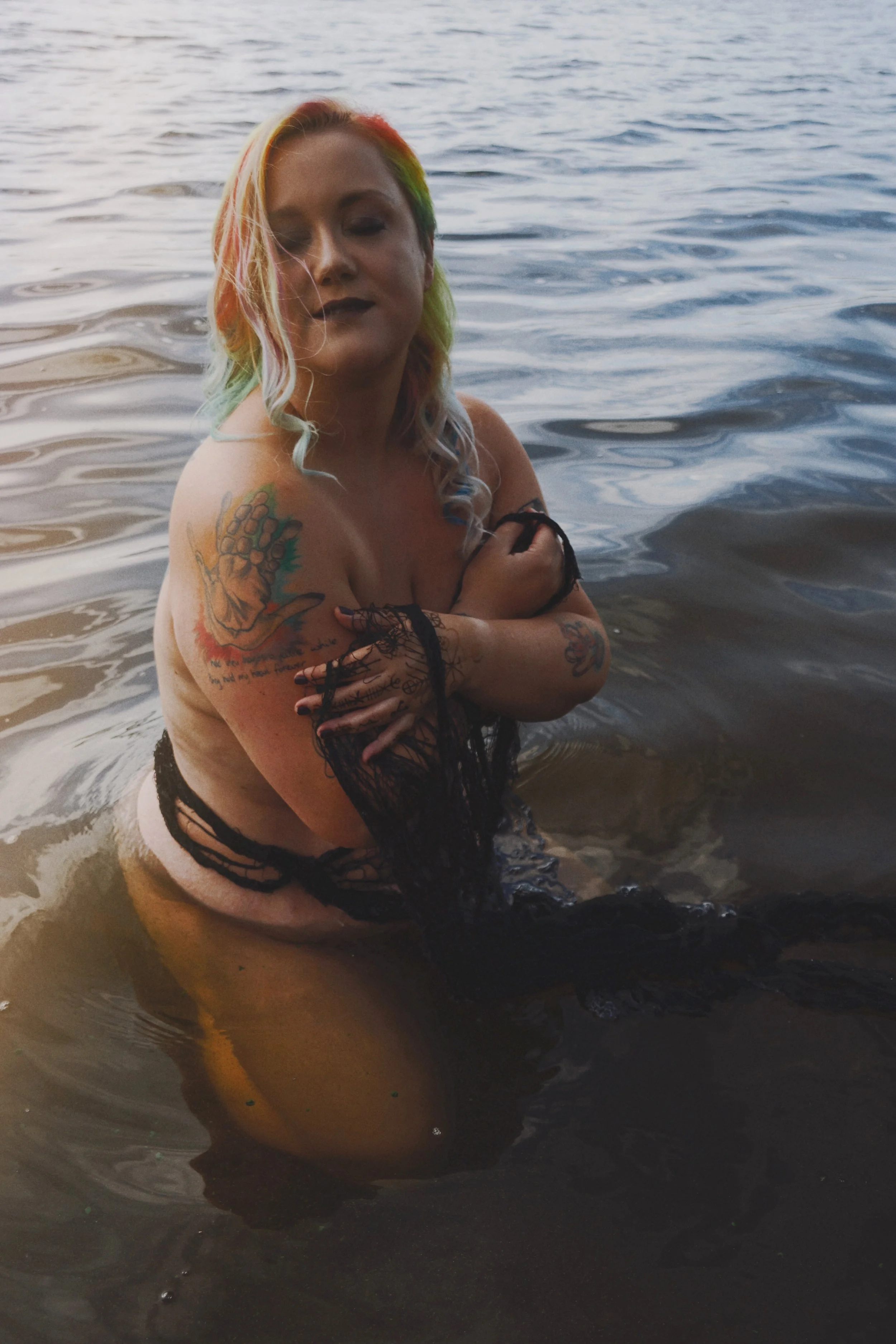 A woman with colorful rainbow hair and tattoos is sitting in a body of water, wearing a black lace garment and looking at the camera with a relaxed expression.
