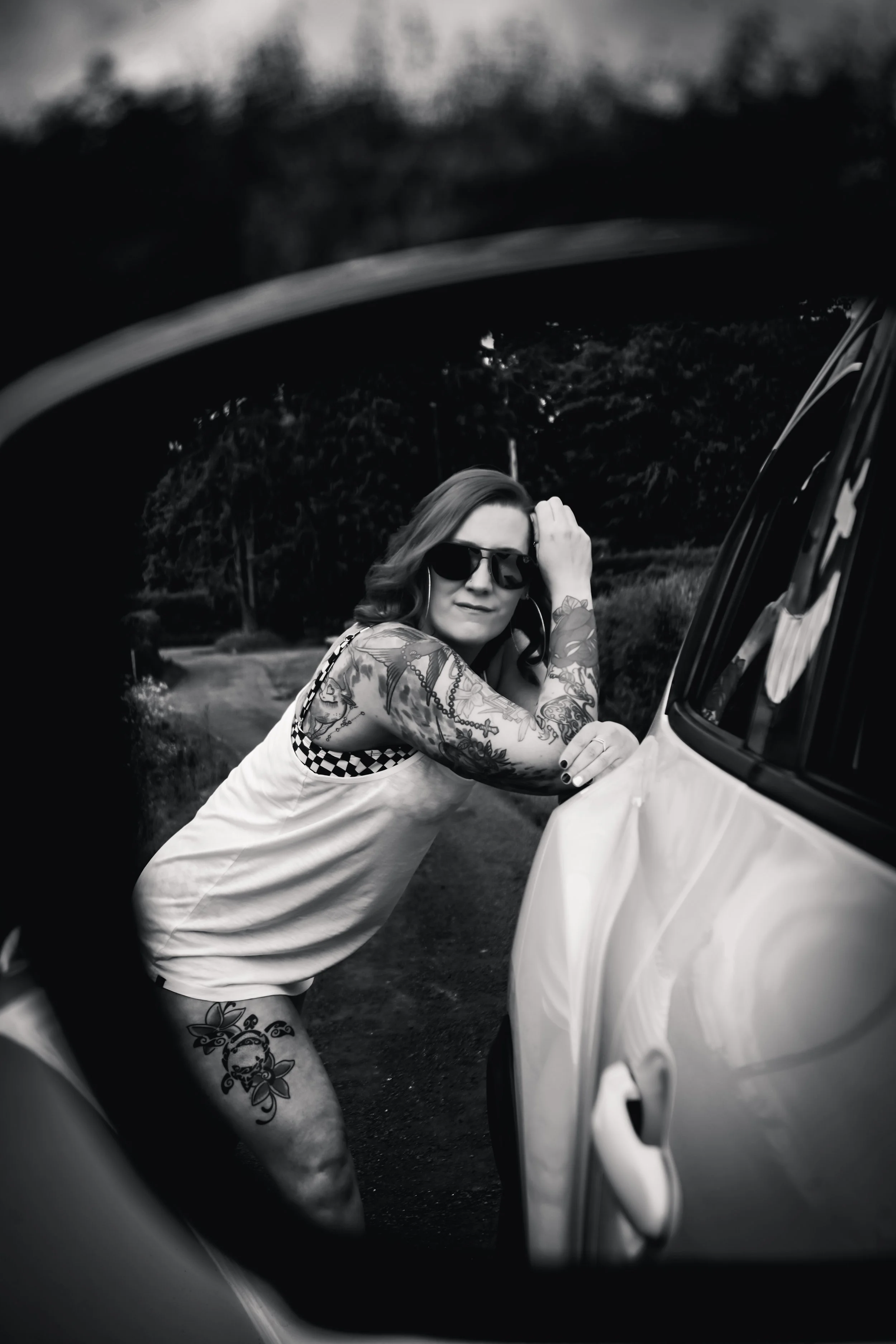 Black and white photo of a woman with tattoos and sunglasses leaning on the side of a car, outdoors on a winding road surrounded by trees.