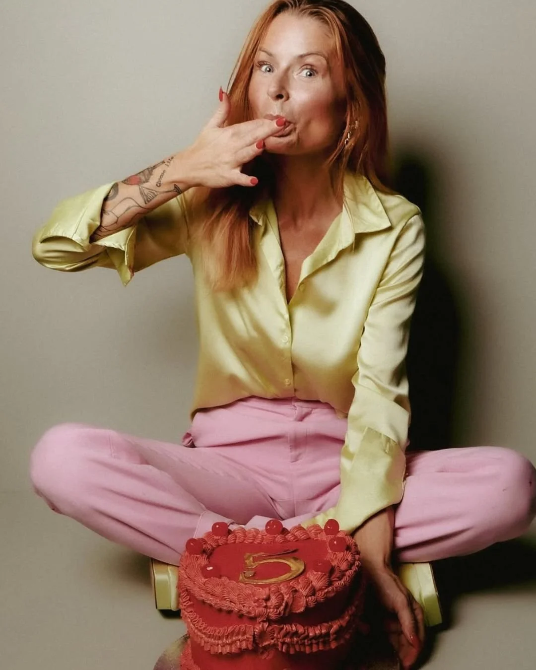 A woman with reddish hair, wearing a yellow satin shirt and pink pants, is sitting cross-legged. She is blowing a kiss with her right hand, which has tattoos, and has red nail polish. There is a red heart-shaped cake with a gold number five candle in front of her on the floor.