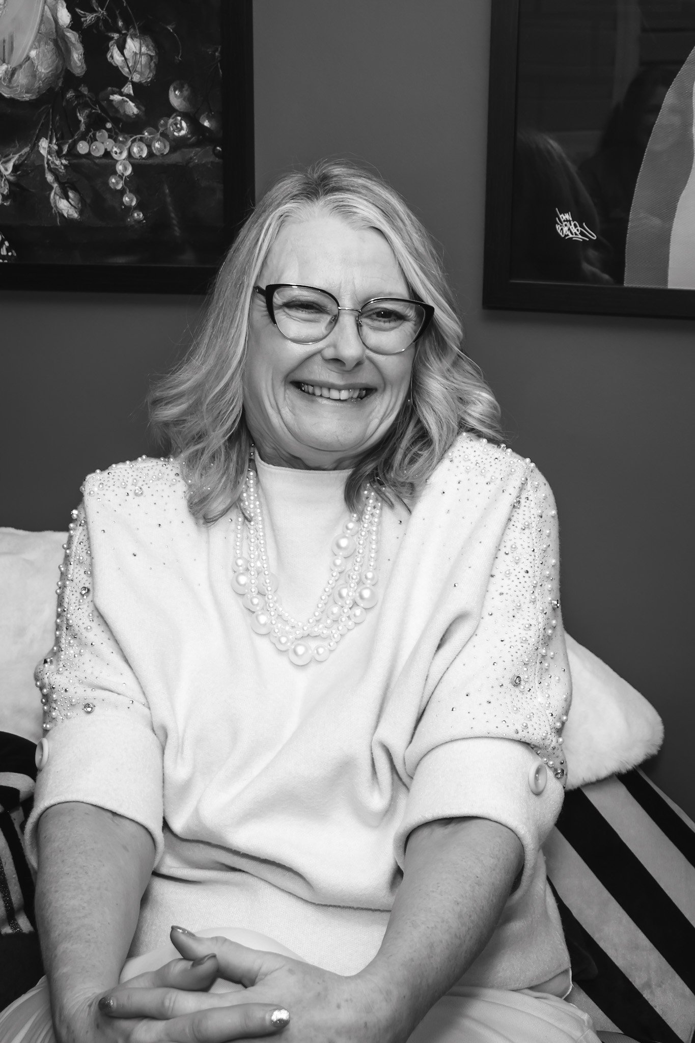 A smiling woman with blonde hair, wearing glasses and a pearl necklace, sitting on a couch in a room with framed artwork on the wall.