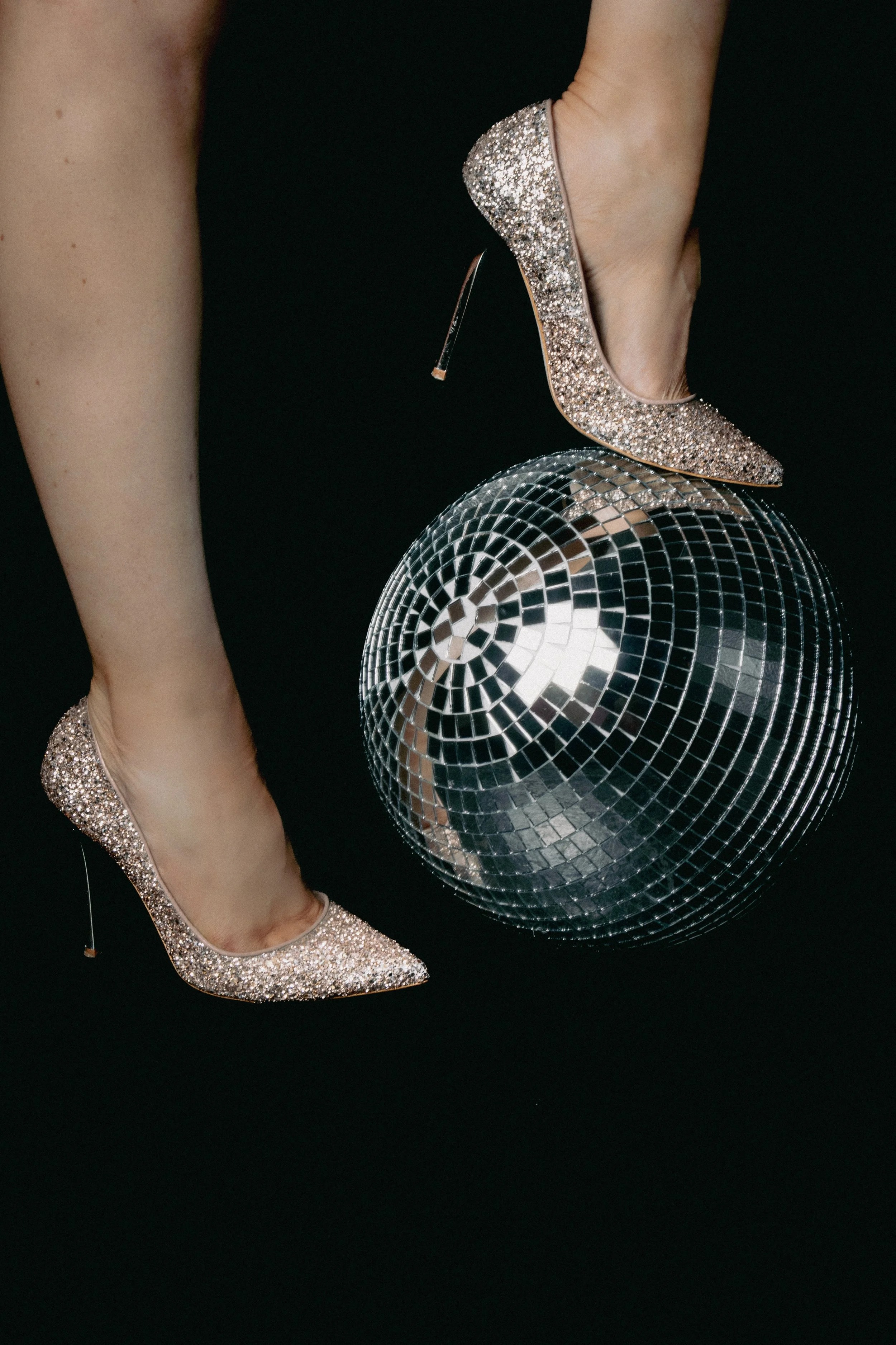 A person wearing glittery high heels balancing on a disco ball, which is on a black background.
