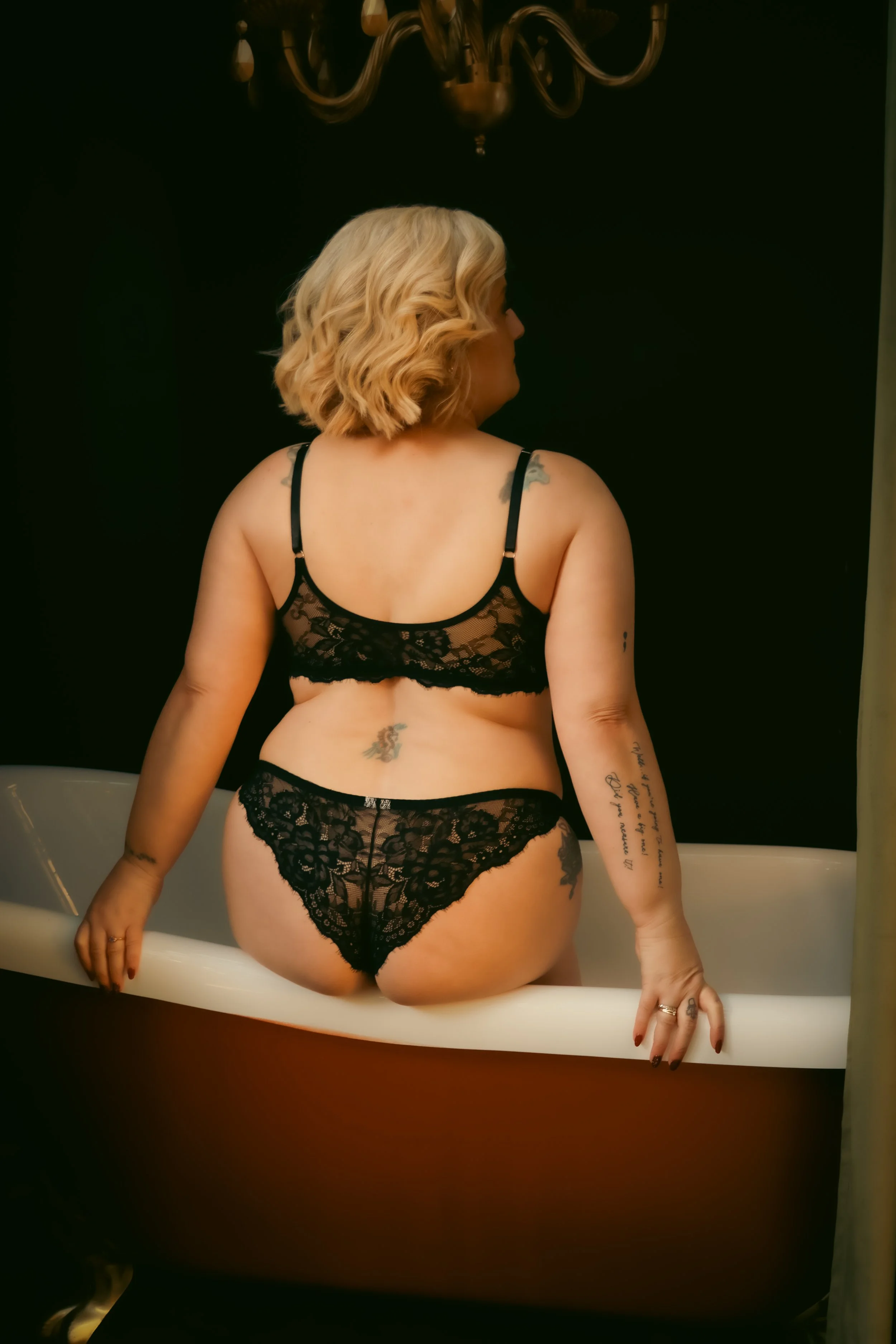 A woman with blonde, wavy hair sitting in a bathtub, dressed in black lace lingerie, with tattoos visible on her back and arms, against a dark background with a chandelier hanging above.