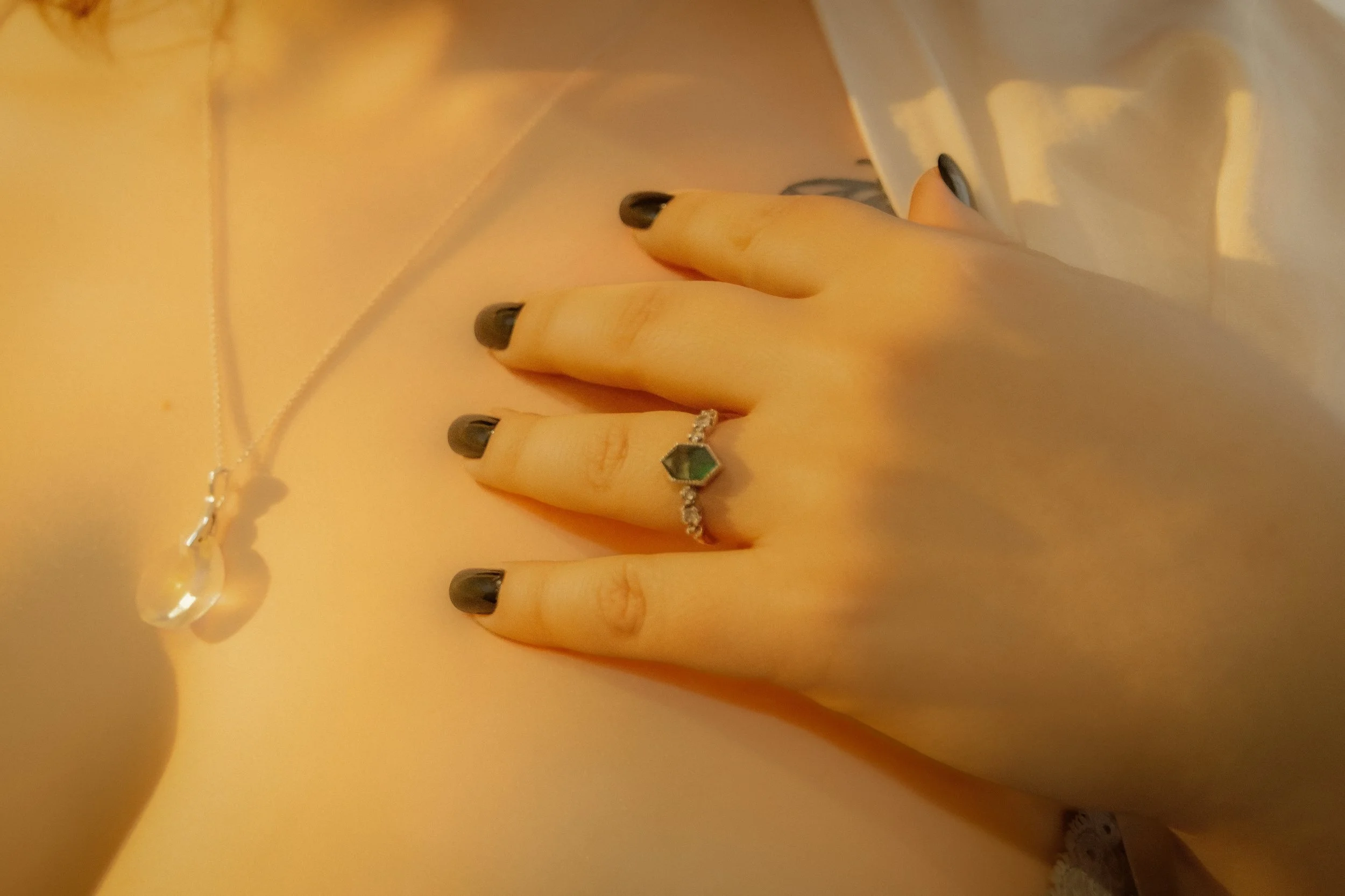 Hand resting on chest wearing a ring with green hexagon-shaped stone and black nail polish, with a delicate necklace featuring a large teardrop-shaped pendant.