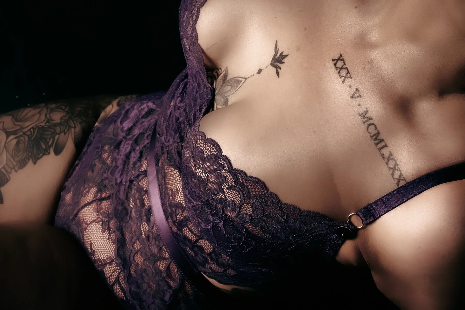 Close-up of a woman's chest and shoulder, wearing purple lace lingerie, showing tattoos on her chest and arm.