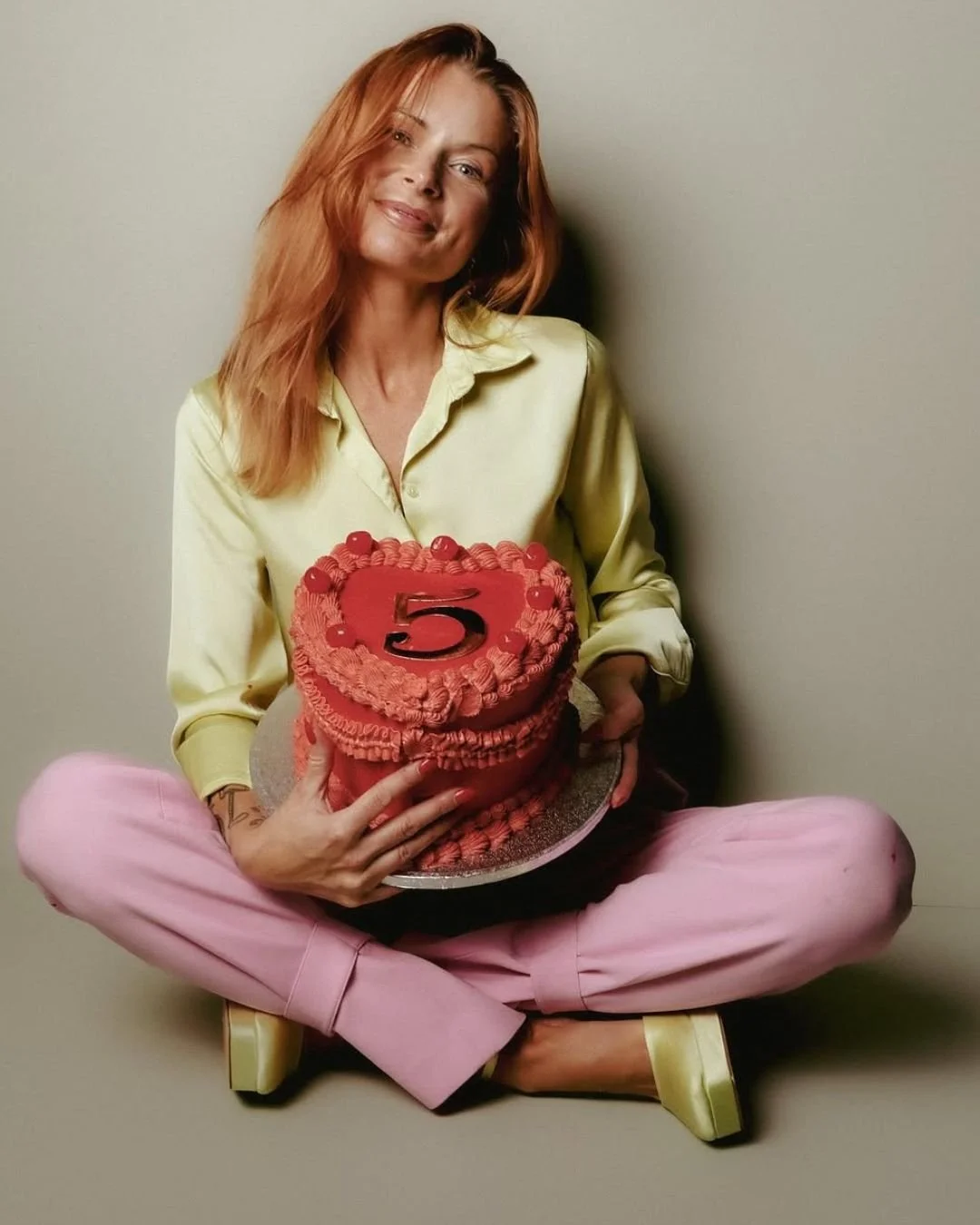 Woman with red hair sitting cross-legged on the floor, holding a red birthday cake with a large number 5 on top.