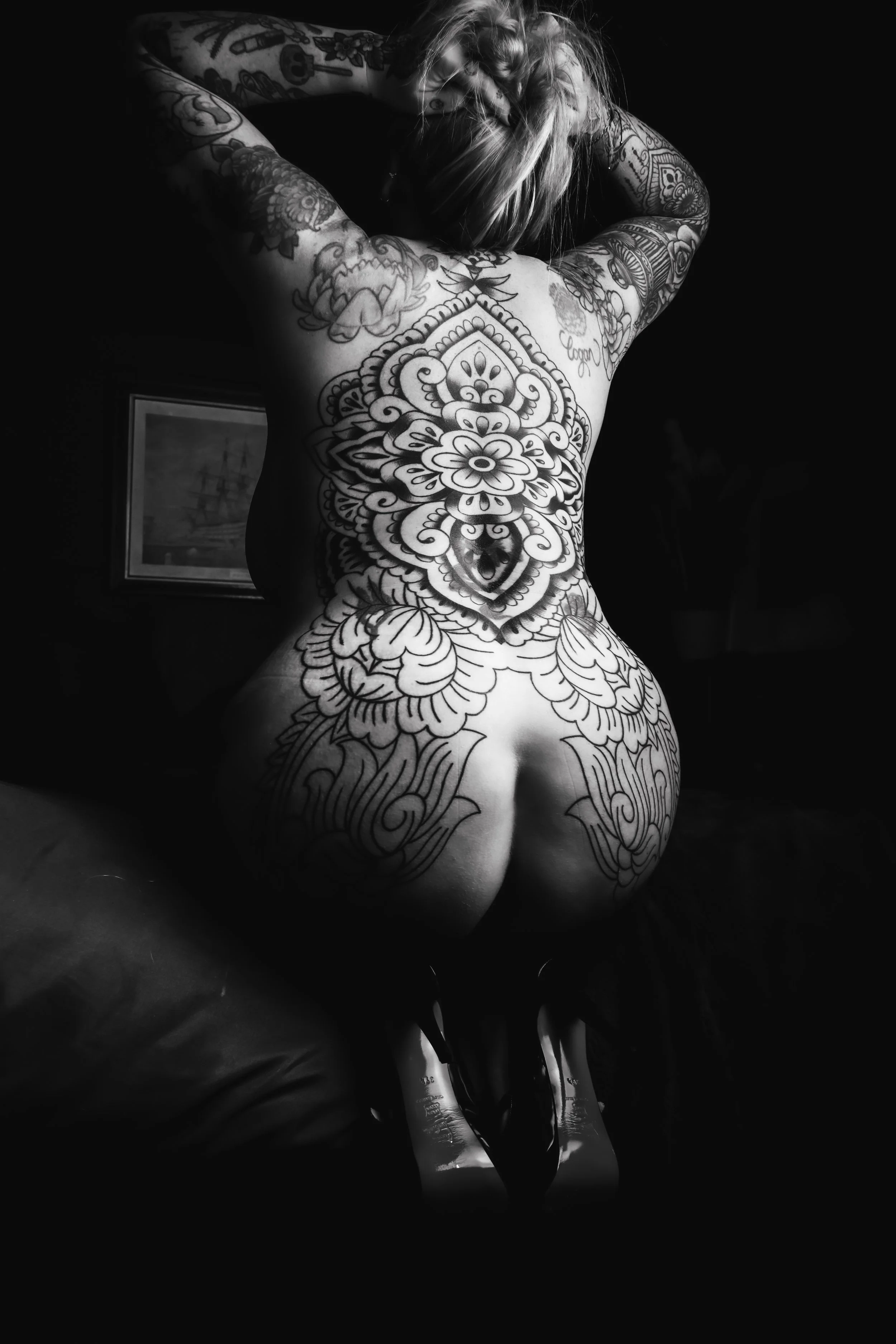 Back view of a tattooed woman with tattoos covering her back, arms, and legs, kneeling with her arms raised and her head resting on her hands, wearing high-heeled shoes.